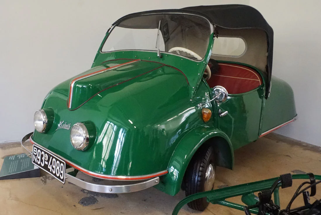 Ein grünes, dreirädriges Auto mit einem Stoffdach und einem runden Heck. Es hat zwei Frontscheinwerfer und eine geschwungene Form. Die Innenausstattung ist rot und das Fahrzeug steht in einem Ausstellungsraum. Das Kennzeichen ist sichtbar: B-N 93-4989.