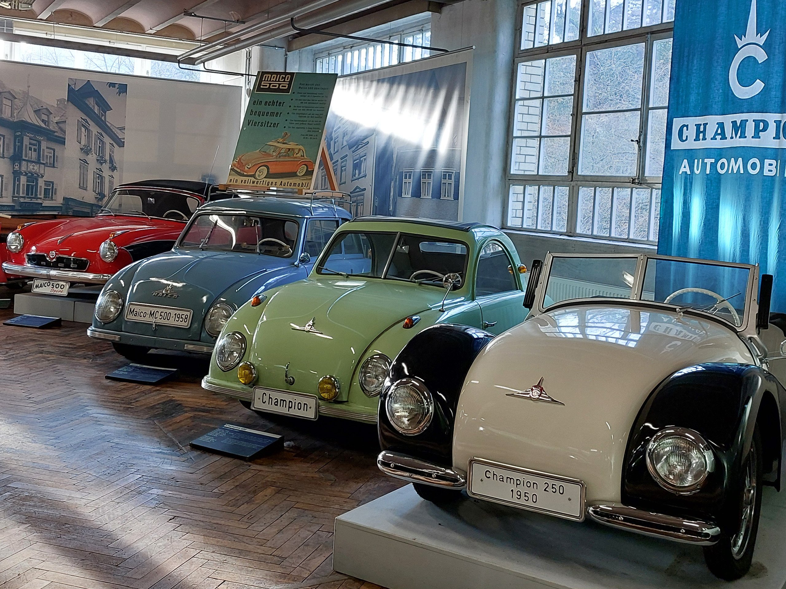 Im Bild sind vier klassische Automobile in einem Ausstellungsraum zu sehen. Die Autos sind in verschiedenen Farben: rot, blau, grün und weiß/ schwarz. Die Modelle sind mit Schildern versehen, die ihre Marken und Baujahre anzeigen. Im Hintergrund sind Fenster und Banner zu erkennen.