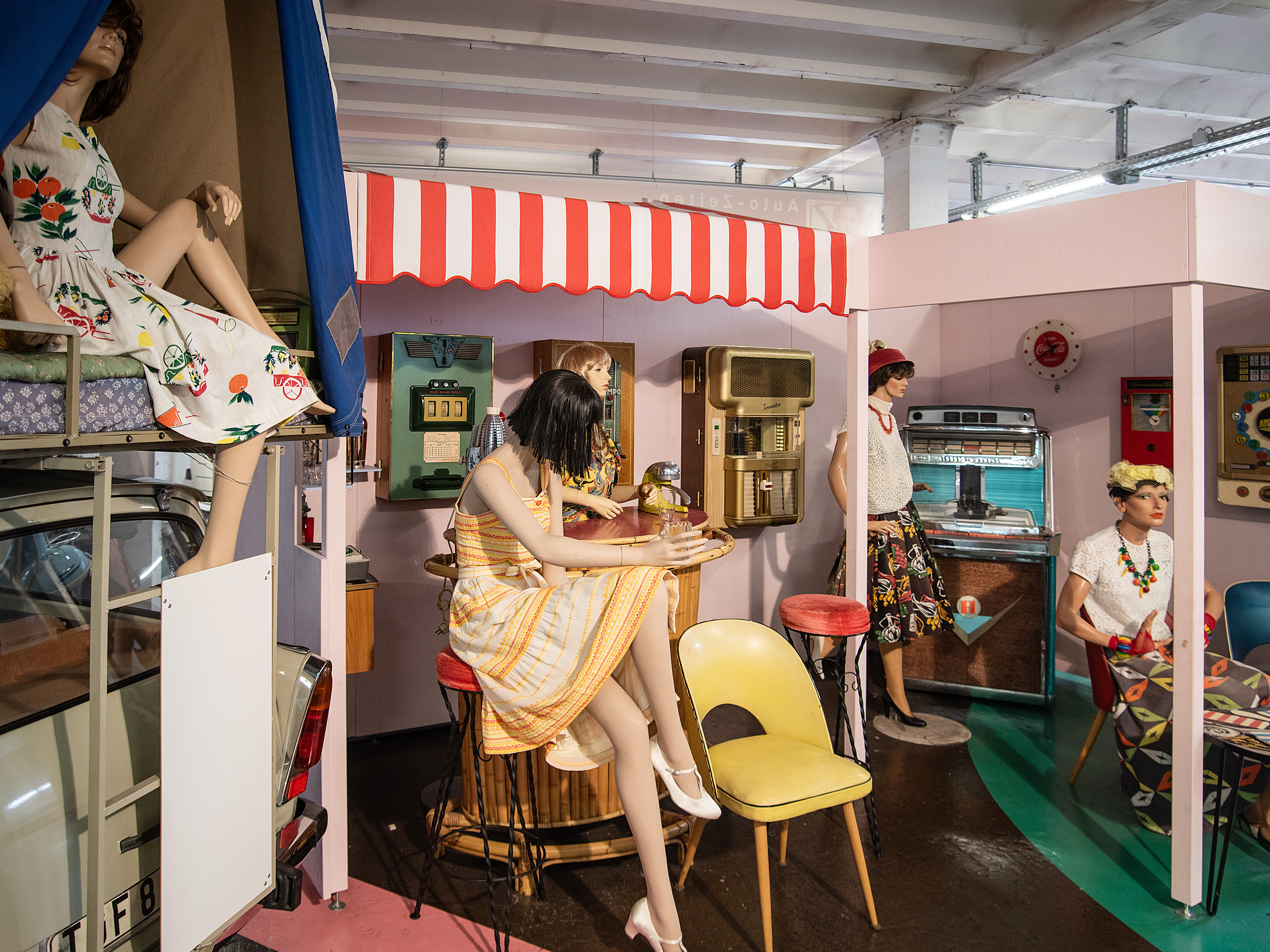 Die Abbildung zeigt eine Ausstellung mit Schaufensterpuppen in vintage-inspirierten Kleidern, die in einem nostalgischen Raum angeordnet sind. Bunte Möbel und Haushaltsgeräte aus vergangener Zeit sind ebenfalls zu sehen, was eine nostalgische Atmosphäre schafft.