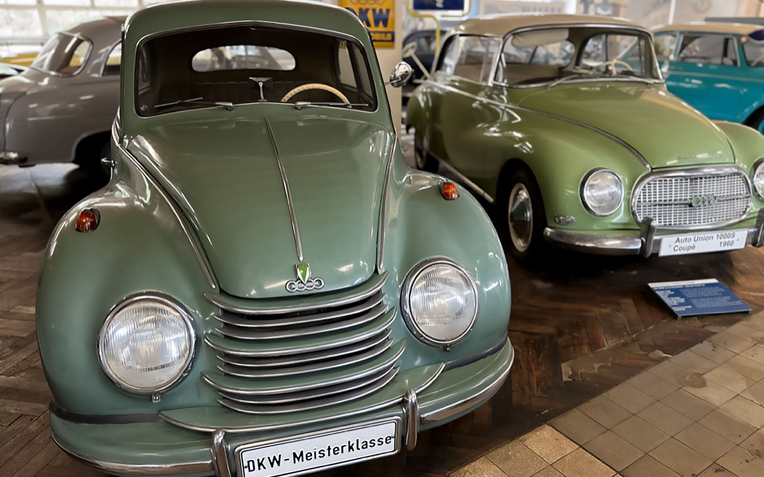 In der Bildmitte steht ein klassischer, mintgrüner Auto mit runder Form und einem schlichten Grill. Daneben ist ein weiterer, hellgrüner Wagen sichtbar. Beide Fahrzeuge sind in einem Automuseum ausgestellt.