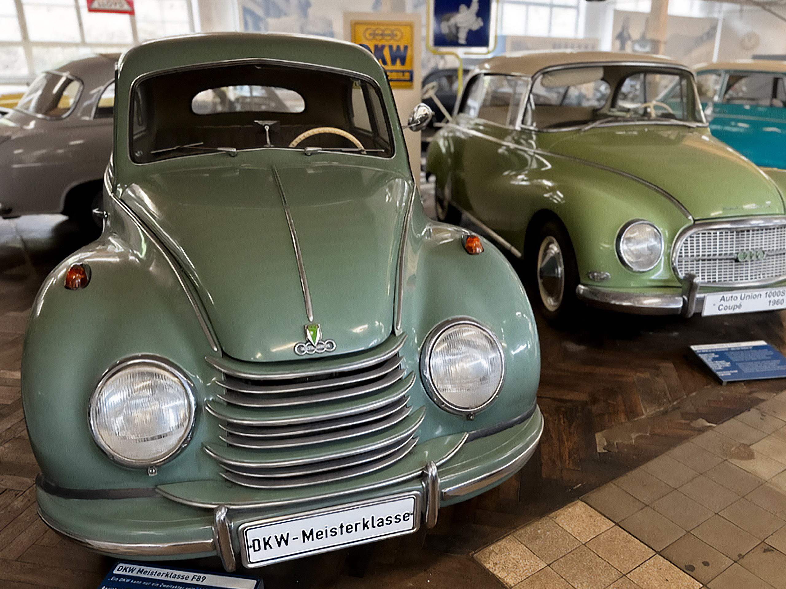 Das Bild zeigt zwei klassische Autos in einem Museum. Im Vordergrund steht ein hellgrüner DKW F89 mit runden Scheinwerfern und einem markanten Kühlergrill. Im Hintergrund ist ein weiteres grünes Coupé zu sehen. Beide Autos repräsentieren Oldtimer-Design.