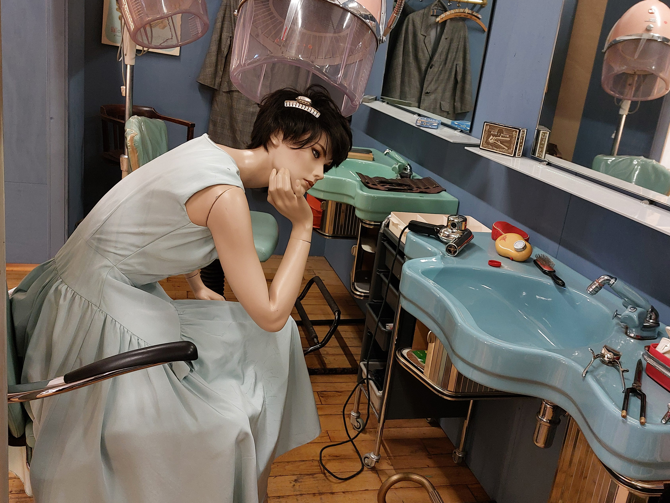 Ein Schaufensterpuppen sitzt in einem Friseursalon in einem mintgrünen Kleid. Sie schaut nachdenklich in einen Spiegel. Im Hintergrund sind Friseurequipment und -stühle zu sehen, die Atmosphäre vermittelt einen nostalgischen Charme.