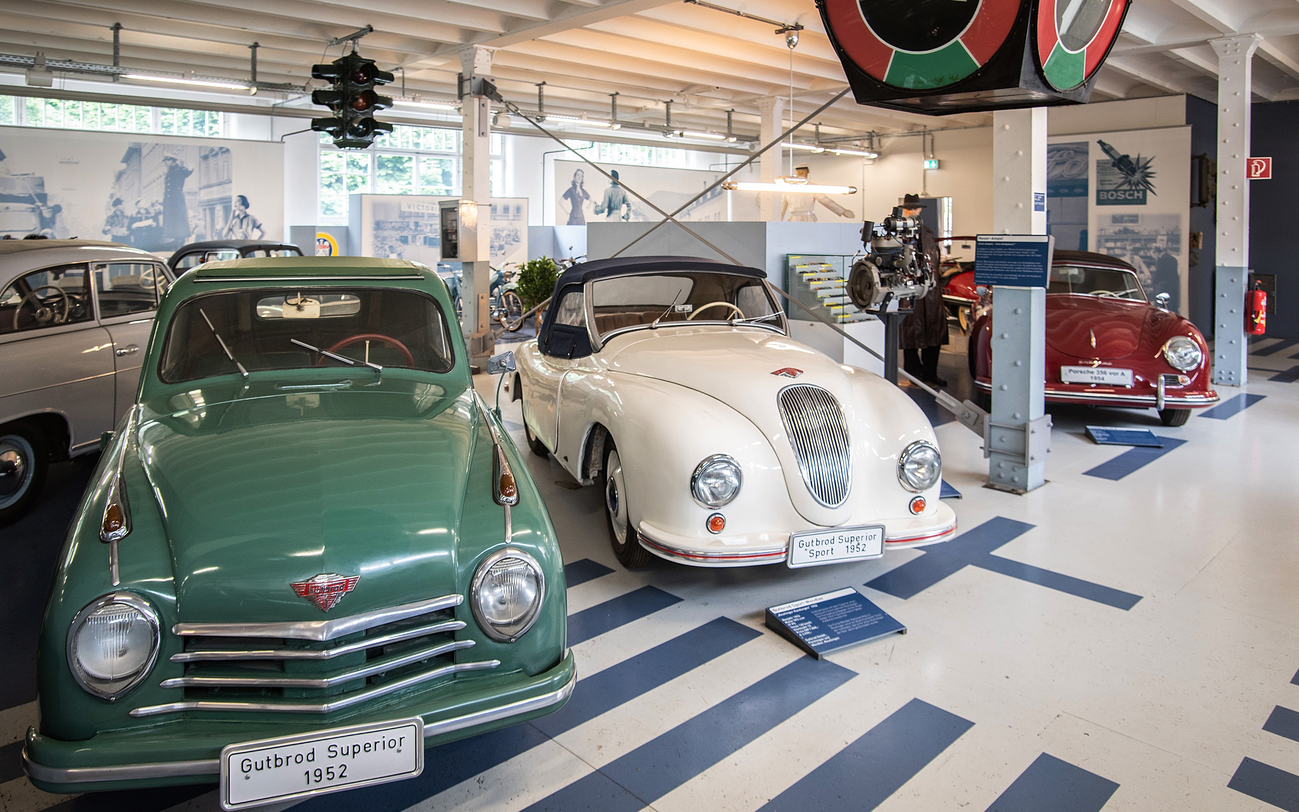 Im Bild sind drei historische Autos in einem Museum zu sehen. Links steht ein grünes Fahrzeug, in der Mitte ein weißes Cabriolet und rechts ein rotes Auto. Die Autos sind auf einem hellen Boden angeordnet, umgeben von Ausstellungsinformationen.