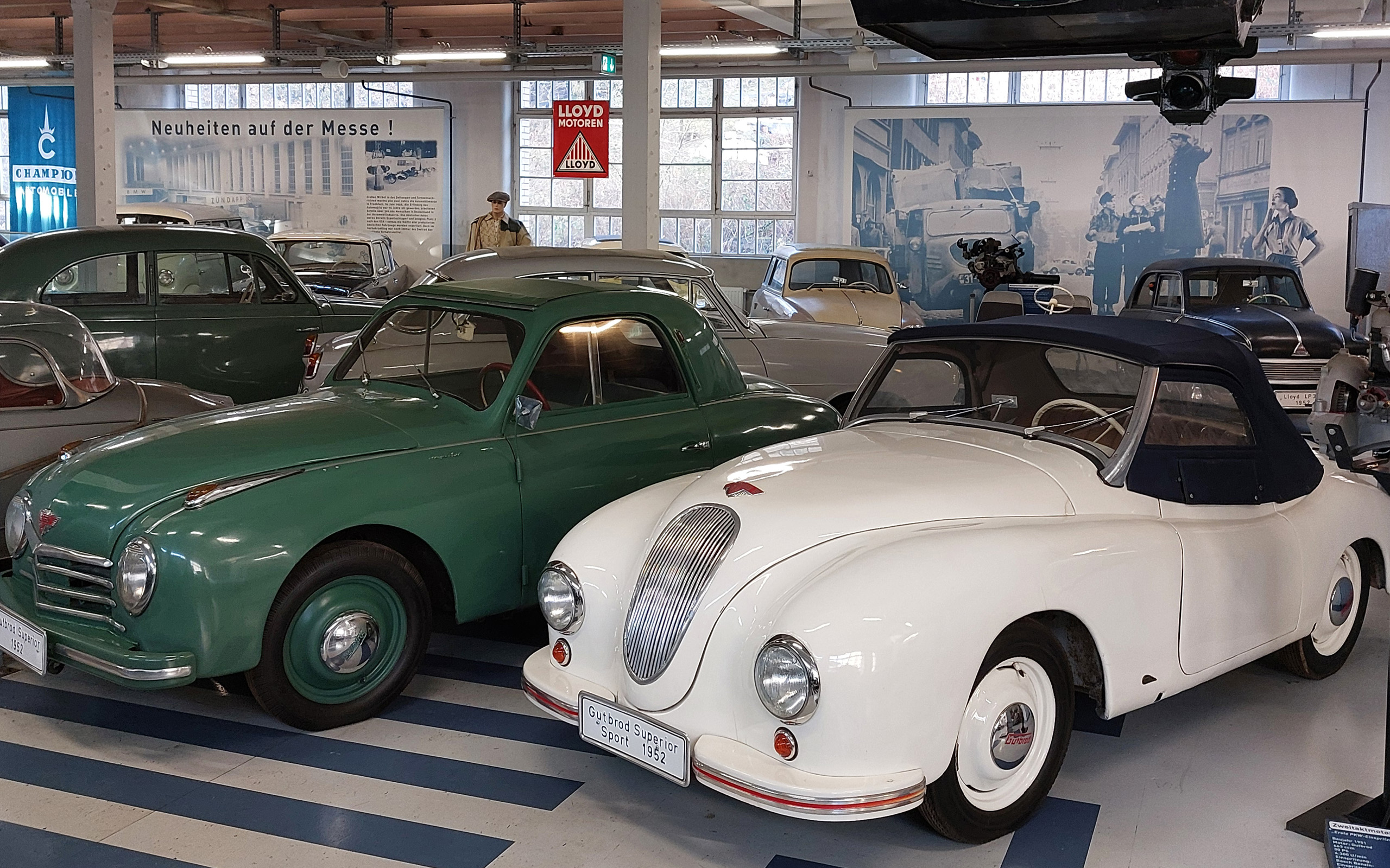 Zwei klassische Autos in einem Ausstellungsraum. Links ein grüner Sportwagen und rechts ein weißes Cabriolet mit einem blauen Verdeck. Im Hintergrund sind weitere historische Fahrzeuge sowie Wände mit Bildern und Schildern sichtbar.