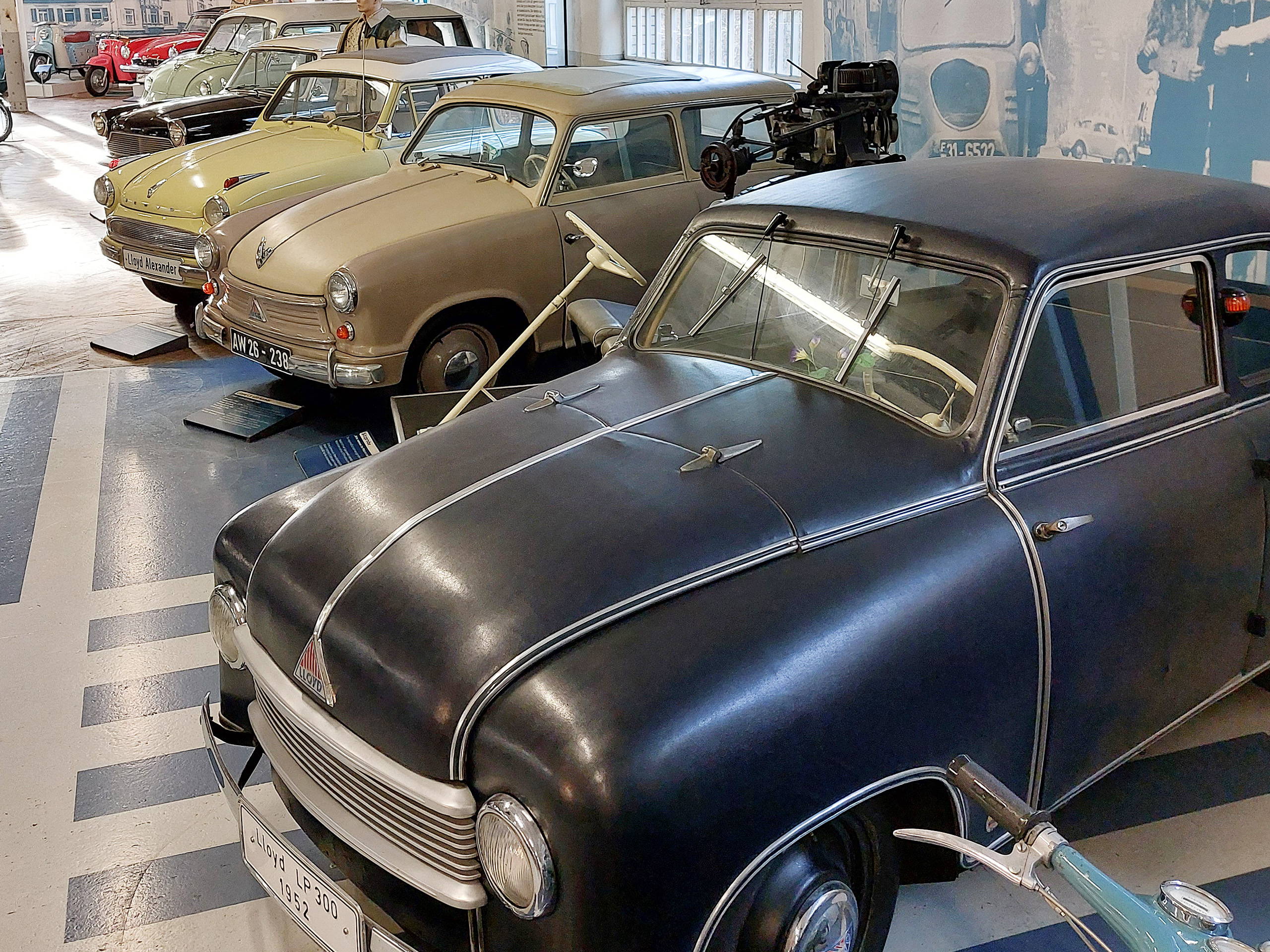 In einem Museum stehen mehrere alte Automobile nebeneinander. Im Vordergrund ist ein schwarzer Lloyd mit auffälligen, geschwungenen Linien zu sehen. Daneben sind Fahrzeuge in Gelb und Braun aus verschiedenen Epochen arrangiert. Im Hintergrund erkennt man einen Ausstellungsbereich mit weiteren Klassikern.