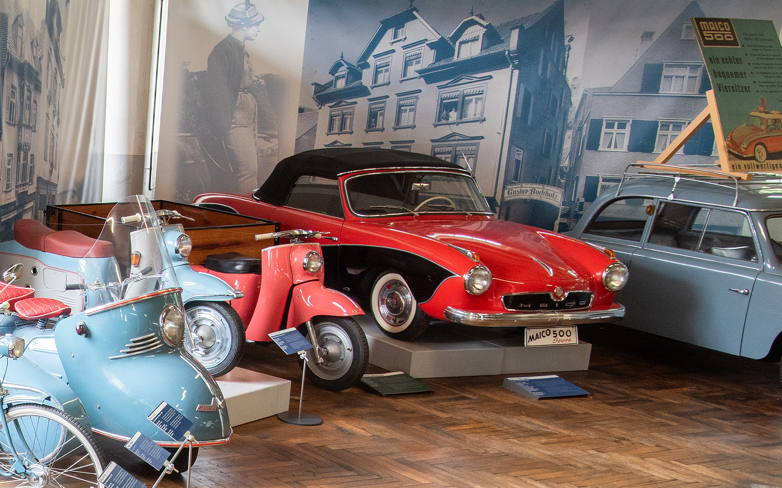Im Bild sind mehrere historische Fahrzeuge in einem Museum zu sehen. Im Vordergrund steht ein rot-schwarzes Auto, ein Maico 500. Daneben stehen zwei blaue Kleinkrafträder. Die Umgebung ist mit alten Fotografien und Informationen über die Fahrzeuge dekoriert.