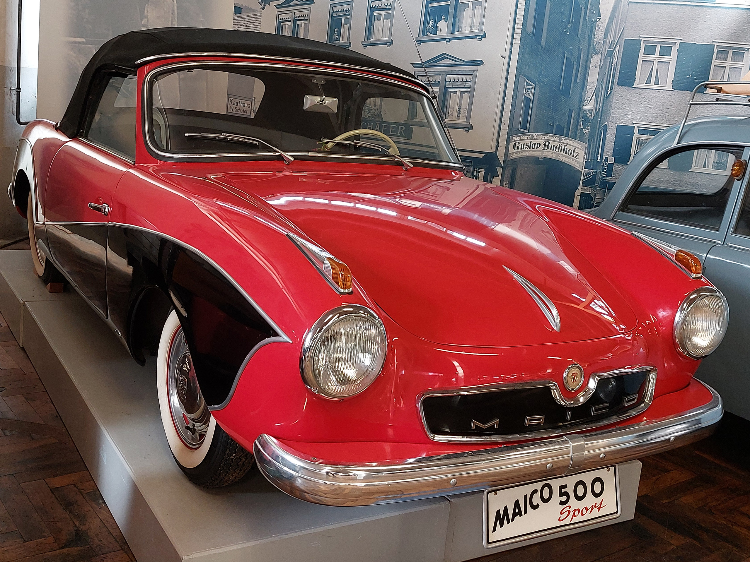 Ein roter Sportwagen mit schwarzem Dach, dem Maico 500, steht auf einem Podest. Der Wagen hat runde Scheinwerfer und einen auffälligen Kühlergrill mit dem Schriftzug "Maico". Im Hintergrund sind unscharfe historische Gebäude zu sehen.