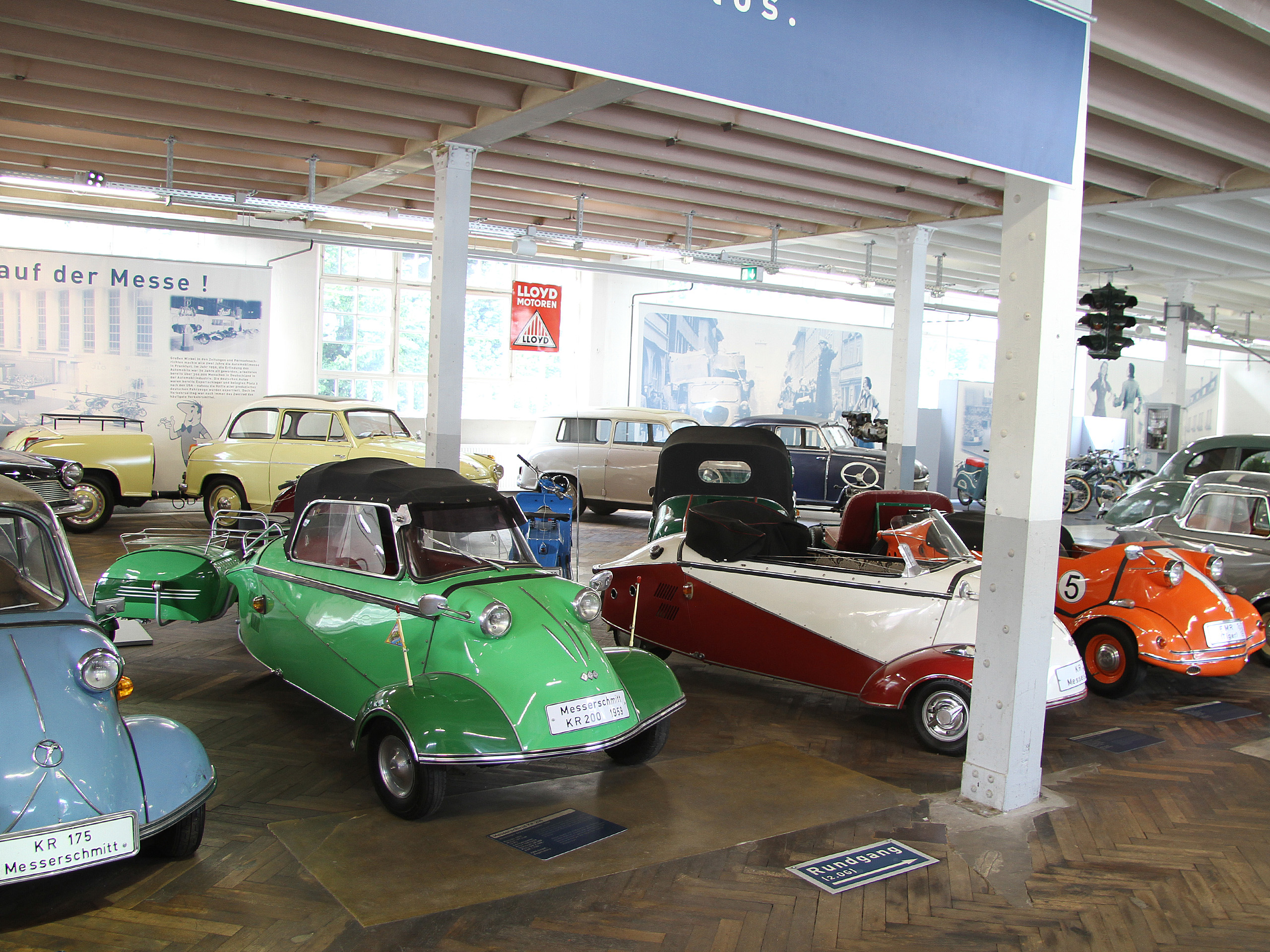 Das Bild zeigt eine Ausstellung mit mehreren bunten Kleinwagen, darunter ein grüner Messerschmitt mit einem schwarzen Verdeck, ein rot-weißer Automobil und ein orangefarbener Wagen. Im Hintergrund sind weitere klassische Autos und historische Informationen sichtbar.
