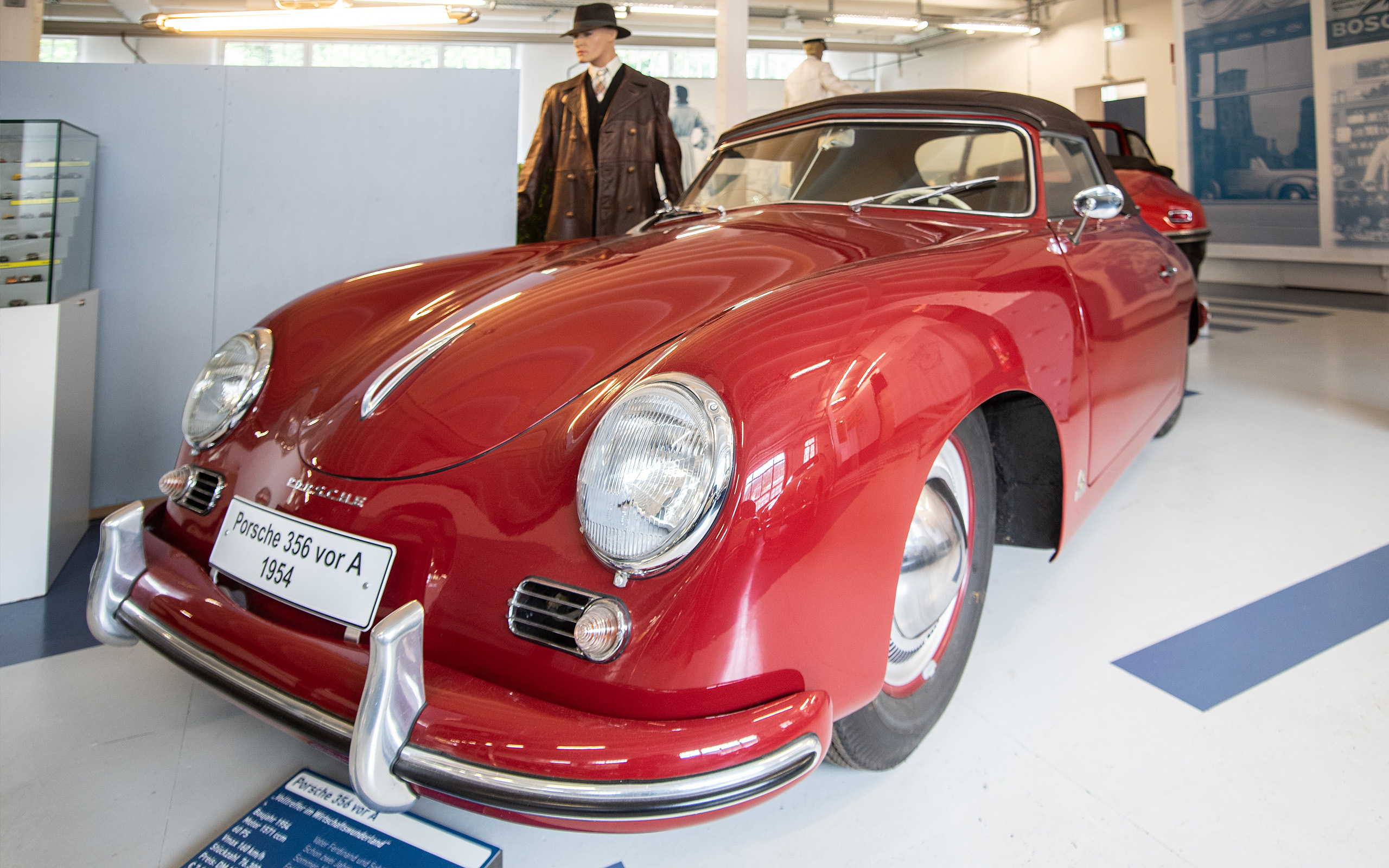Ein roter Porsche 356 vor A aus dem Jahr 1954 steht in einem Ausstellungsraum. Das Auto hat runde Scheinwerfer und eine geschwungene Karosserie. Im Hintergrund sind weitere Fahrzeuge und Ausstellungsstücke zu erkennen.