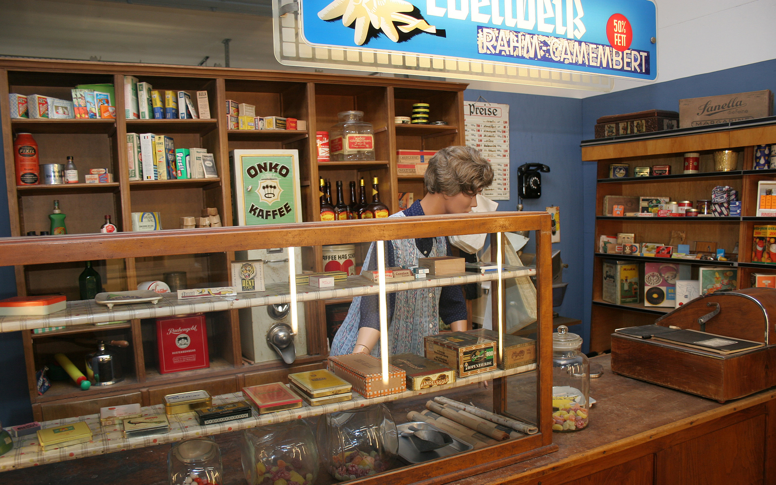 Das Bild zeigt eine nostalgische Verkaufsstelle mit einem Holzregal voller Lebensmittelverpackungen und Flaschen. Im Vordergrund steht eine Person mit grauen Haaren, die hinter einem Glasdisplay arbeitet, das Süßigkeiten und Konservendosen zeigt. Ein Schild mit der Aufschrift "ONKO KAFFEE" ist gut sichtbar.