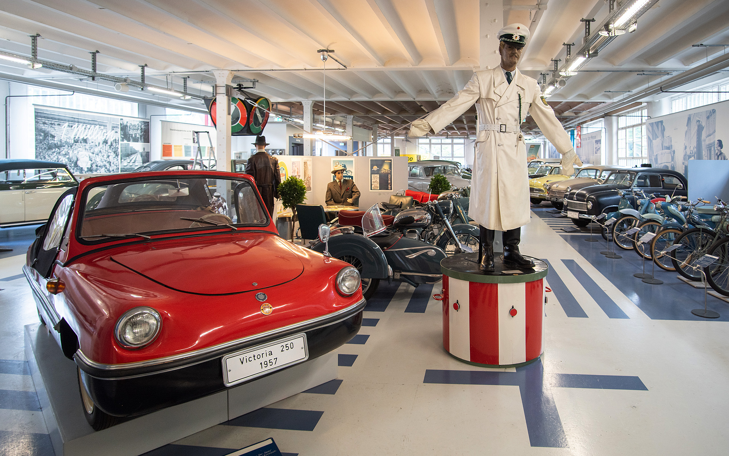 In einem Automuseum steht ein rotes Auto aus dem Jahr 1957 mit einer Beschriftung „Victoria 250“. Neben dem Auto befindet sich eine Figur in einer weißen Uniform, die auf einem Podest steht. Im Hintergrund sind weitere Oldtimer und Motorräder zu sehen.