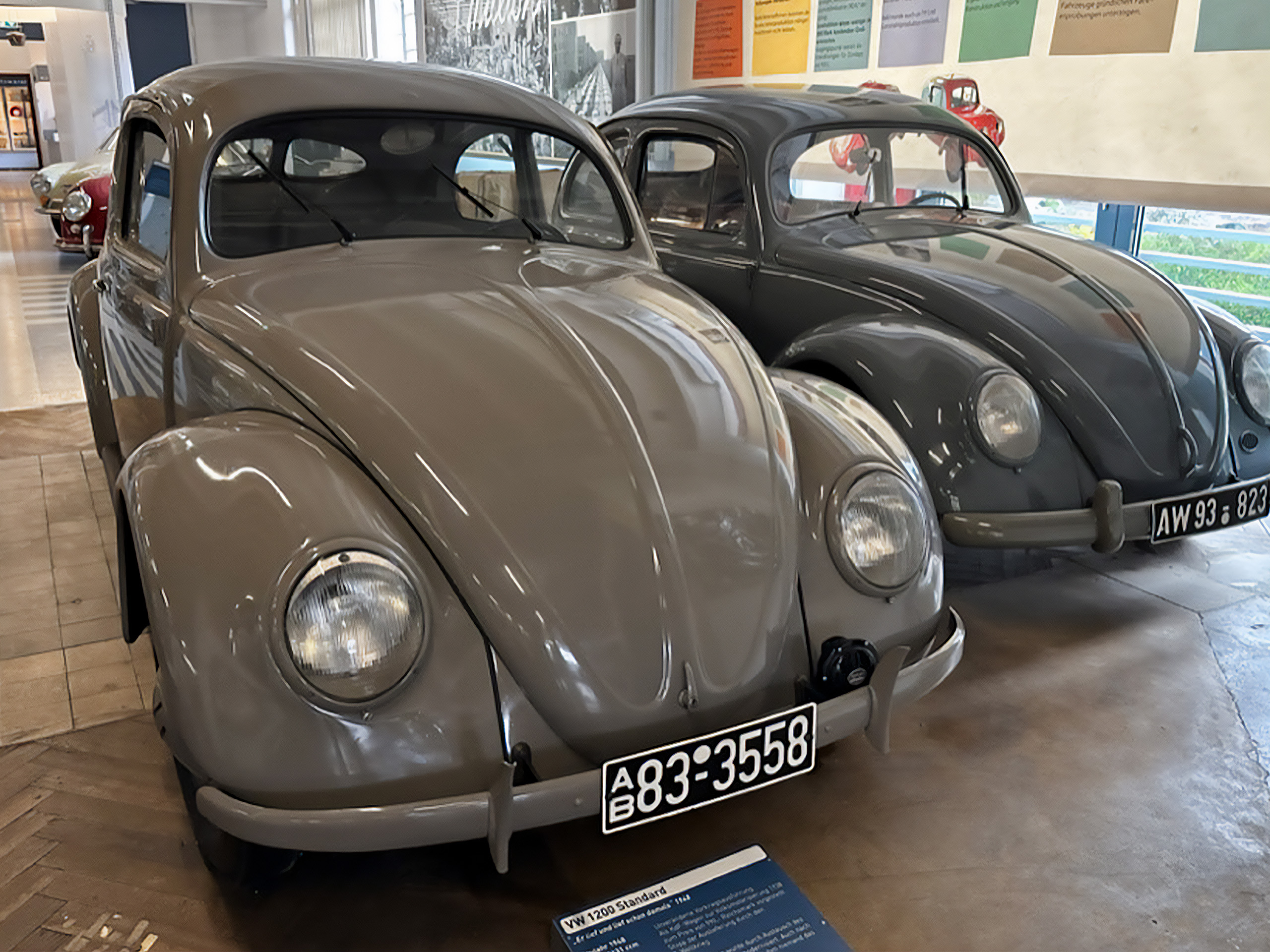 Das Bild zeigt zwei Oldtimer-Fahrzeuge, die nebeneinander in einem Ausstellungsraum stehen. Der linke Wagen ist grau mit einer klassischen Form und einer runden Front. Beide Autos haben runde Scheinwerfer und sind in gutem Zustand.