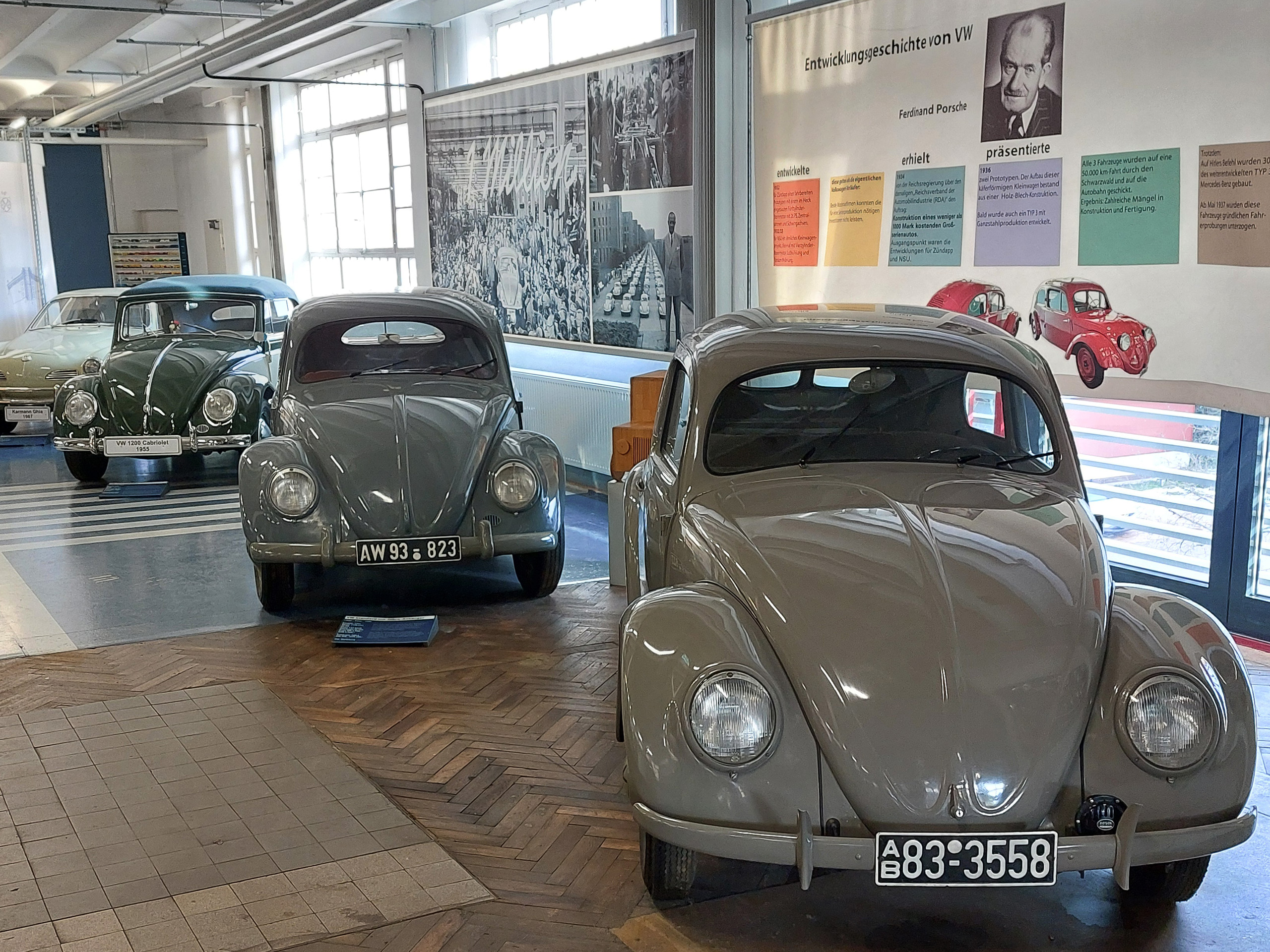 Das Bild zeigt mehrere klassische Volkswagen Käfer in einem Museum. Der zentral platzierte graue Käfer hat die Nummernschilder AW 93 823 und AB 83 3558. Im Hintergrund sind historische Fotos und Informationen zur Entwicklungsgeschichte von VW an der Wand zu sehen.