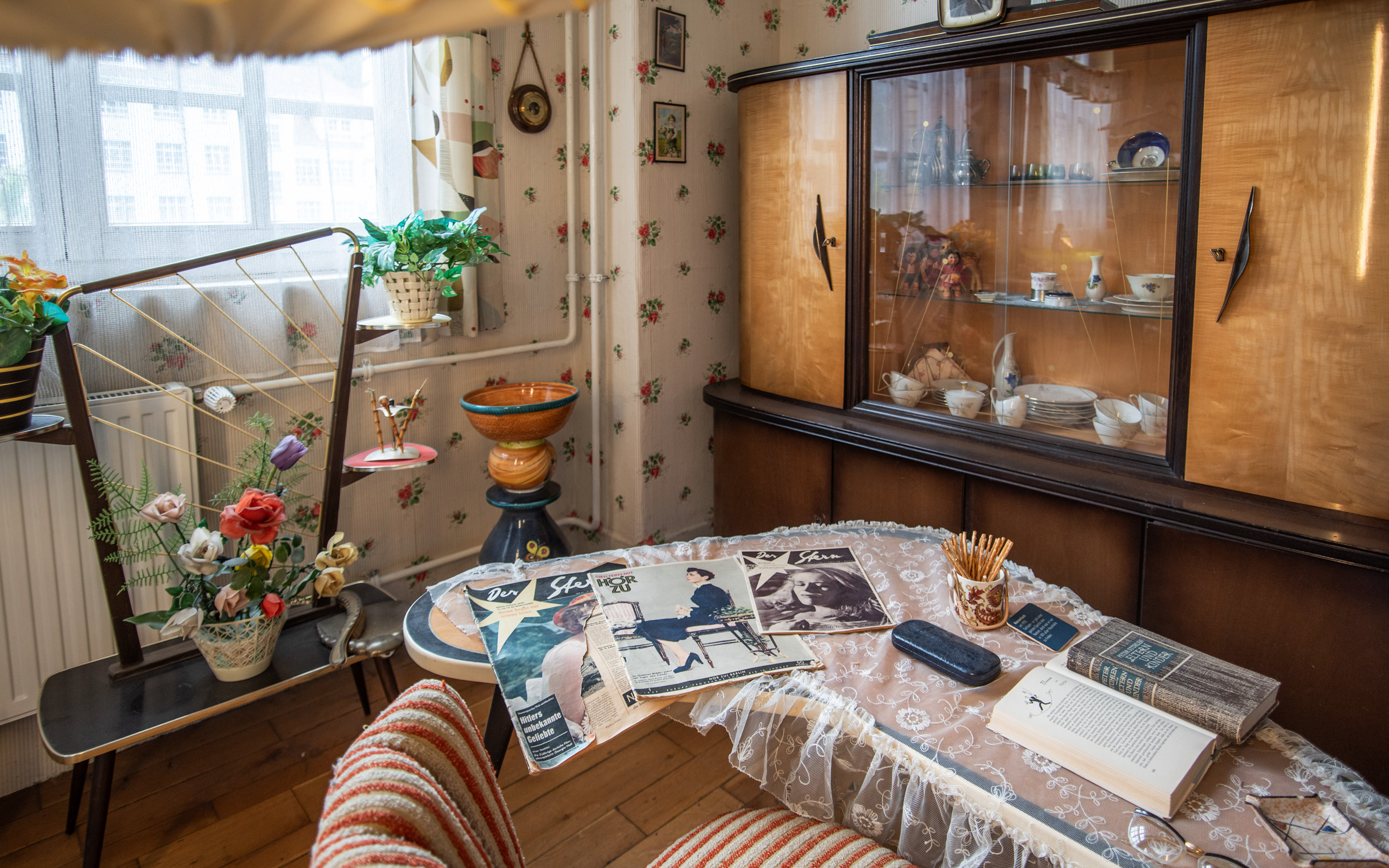 Ein stilvolles Zimmer mit einem Tisch, auf dem Zeitschriften, ein Buch und ein Behälter mit Stiften liegen. Im Hintergrund steht eine Vitrine mit Geschirr, und ein Hocker mit einem dekorativen Topf. Blumen schmücken das Zimmer an einer Wäscheleine. Leichte, helle Vorhänge lassen Tageslicht herein.