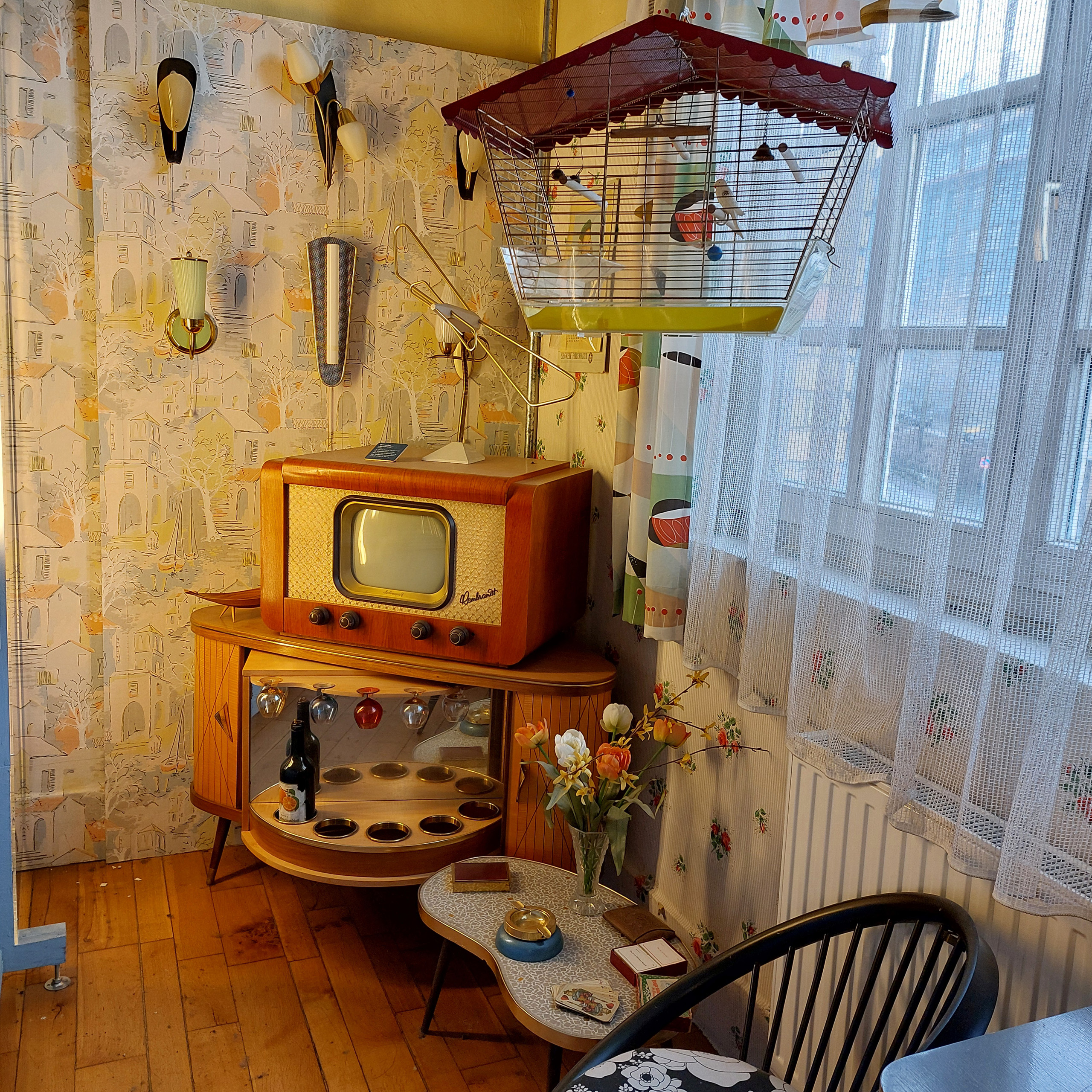 Ein nostalgischer Raum mit einer altmodischen Holzfernsehmaschine, die auf einem niedrigen Tisch steht. Daneben befinden sich ein Blumenstrauß in einer Vase und ein kleiner Tisch mit verschiedenen Gegenständen. Helle Wände mit einer verspielten Tapete und Vorhängen, die Licht hereinlassen.