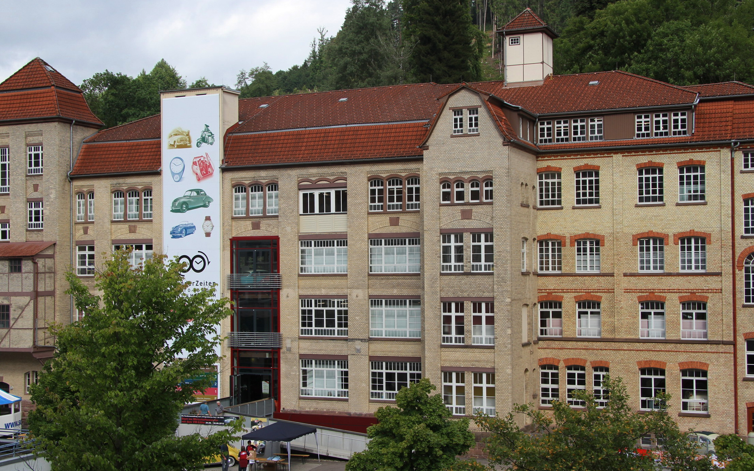 Ein historisches Gebäude mit rotem Ziegeldach und großen Fenstern. An der Vorderseite befindet sich ein großes Plakat mit bunten Illustrationen von Fahrzeugen und Uhren. Im Vordergrund sind Bäume und ein kleiner Gesprächsbereich sichtbar.