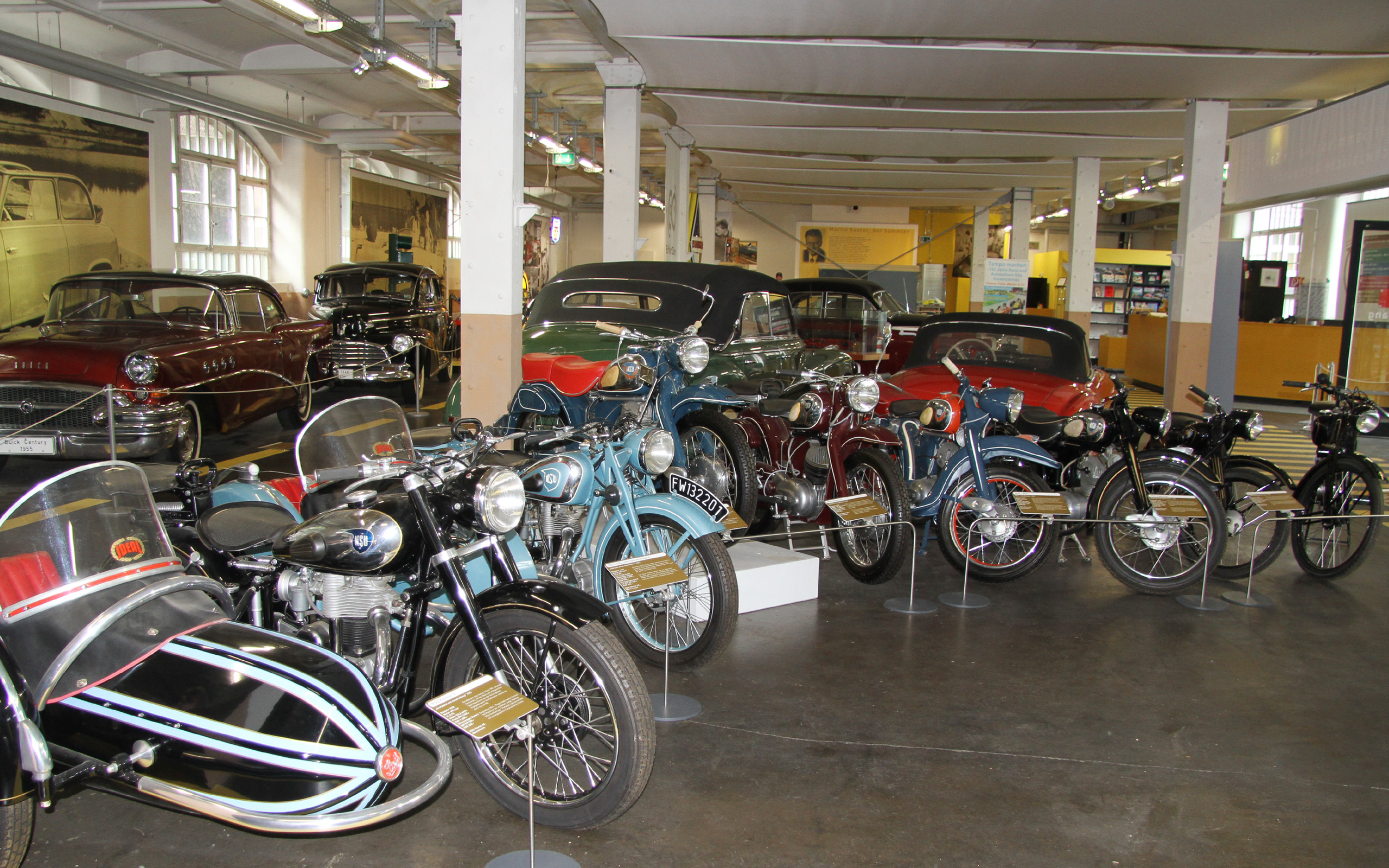 In einem Museum sind mehrere historische Motorräder und Autos ausgestellt. Im Vordergrund stehen Motorräder mit Beiwagen in verschiedenen Farben, darunter blau, schwarz und rot. Im Hintergrund sind klassische Autos zu sehen. Die Umgebung wirkt industriell mit großen Fenstern und Holzböden.
