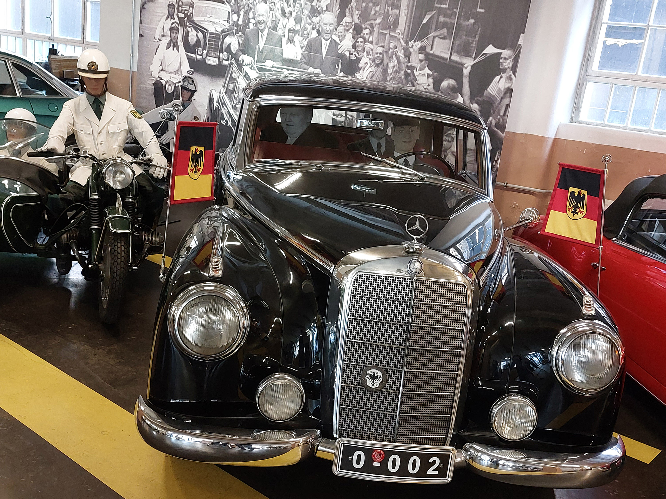Ein schwarzer Mercedes mit eleganter Frontpartie steht in einer Ausstellung. Neben dem Auto steht eine Wachsfigur eines Polizisten auf einem Motorrad mit Beiwagen. Im Hintergrund sind alte Fotos von Fahrzeugen und Menschen zu sehen. Ein Schild mit der deutschen Flagge ziert das Auto.