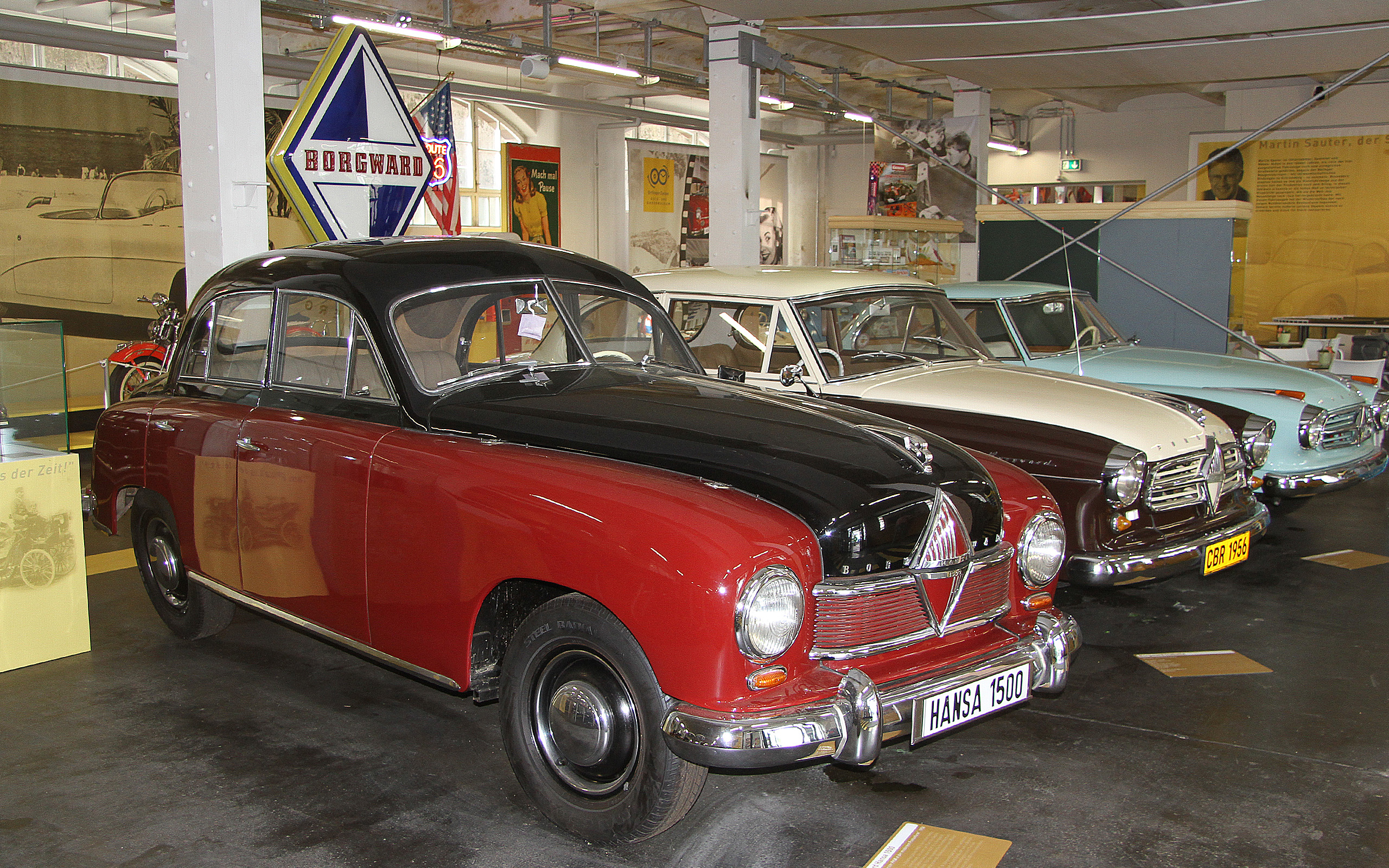 Ein roter und schwarzer Oldtimer steht in einem Automuseum. Im Hintergrund sind weitere klassische Fahrzeuge zu sehen, darunter ein hellblaues und ein beiges Modell. Ein leuchtendes Borgward-Schild ist über den Autos angebracht, was auf die Marke hinweist.