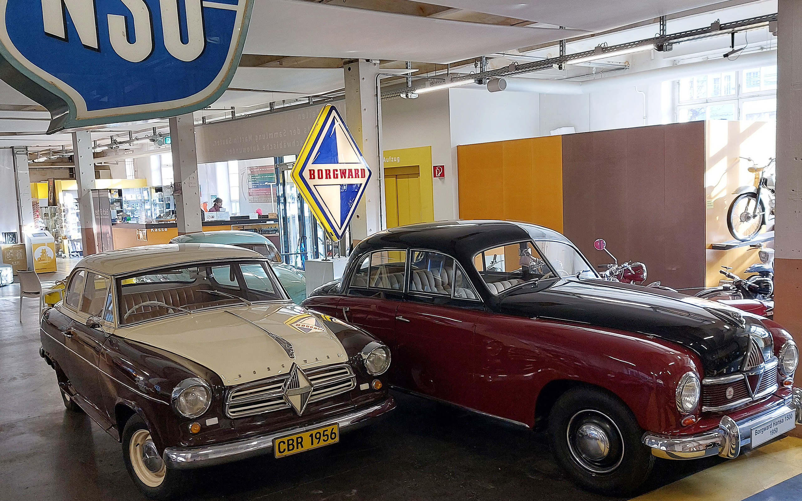 Im Bild sind zwei vintage Autos in einem Ausstellungsraum zu sehen. Das linke Fahrzeug ist ein brauner Wagen mit nostalgischen Details und Nummernschild „CBR 1956“. Das rechte Auto ist rot mit einem schwarzen Dach. Im Hintergrund sind Beschilderungen von NSU und Borgward sichtbar.