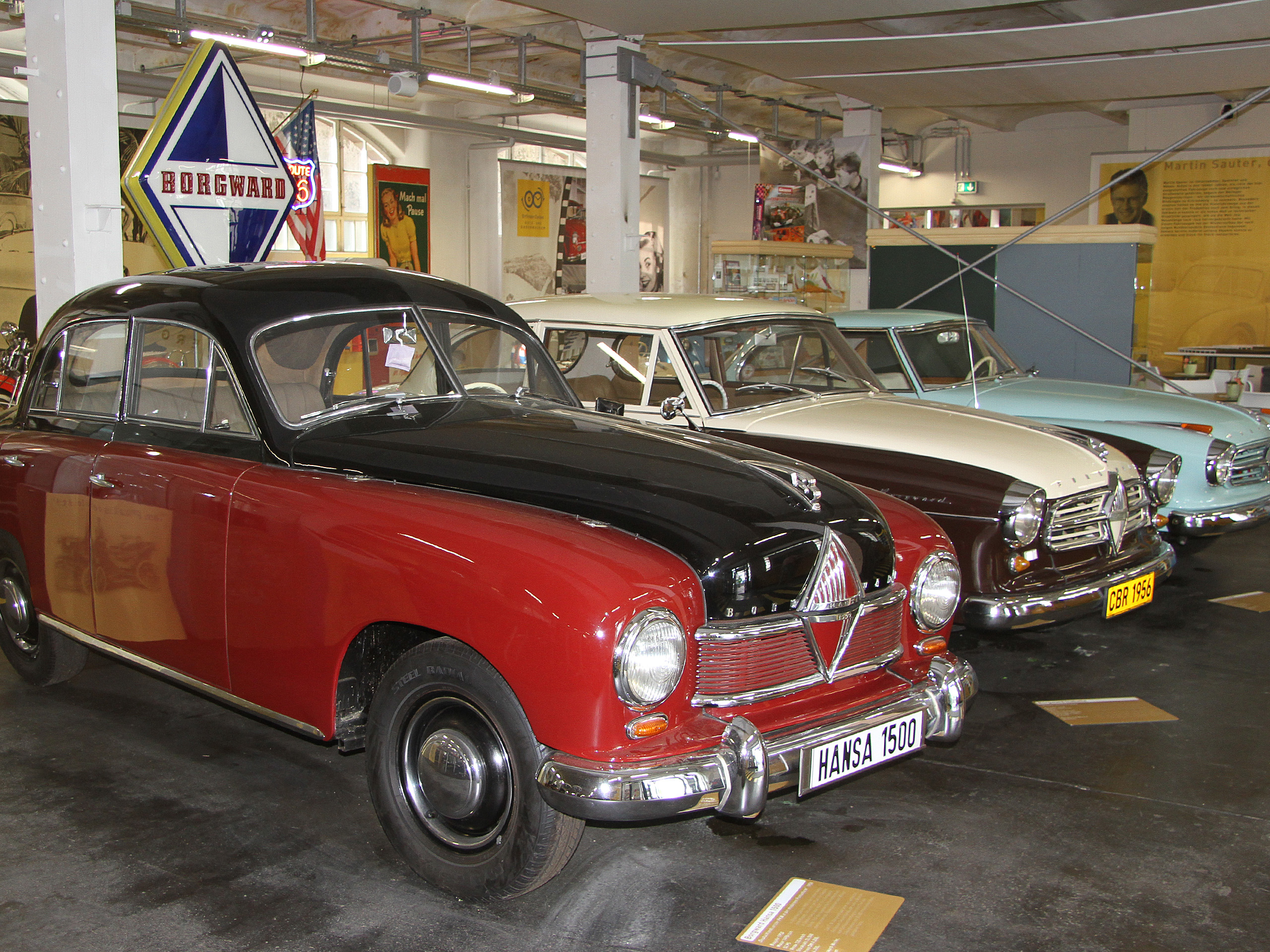 In einem Automuseum sind mehrere klassische Fahrzeuge zu sehen. Im Vordergrund steht ein rot-schwarzes Auto, während im Hintergrund weitere Autos in verschiedenen Farben, einschließlich Weiß und Blau, ausgestellt sind. Ein Schild mit dem Namen Borgward hängt über den Fahrzeugen.