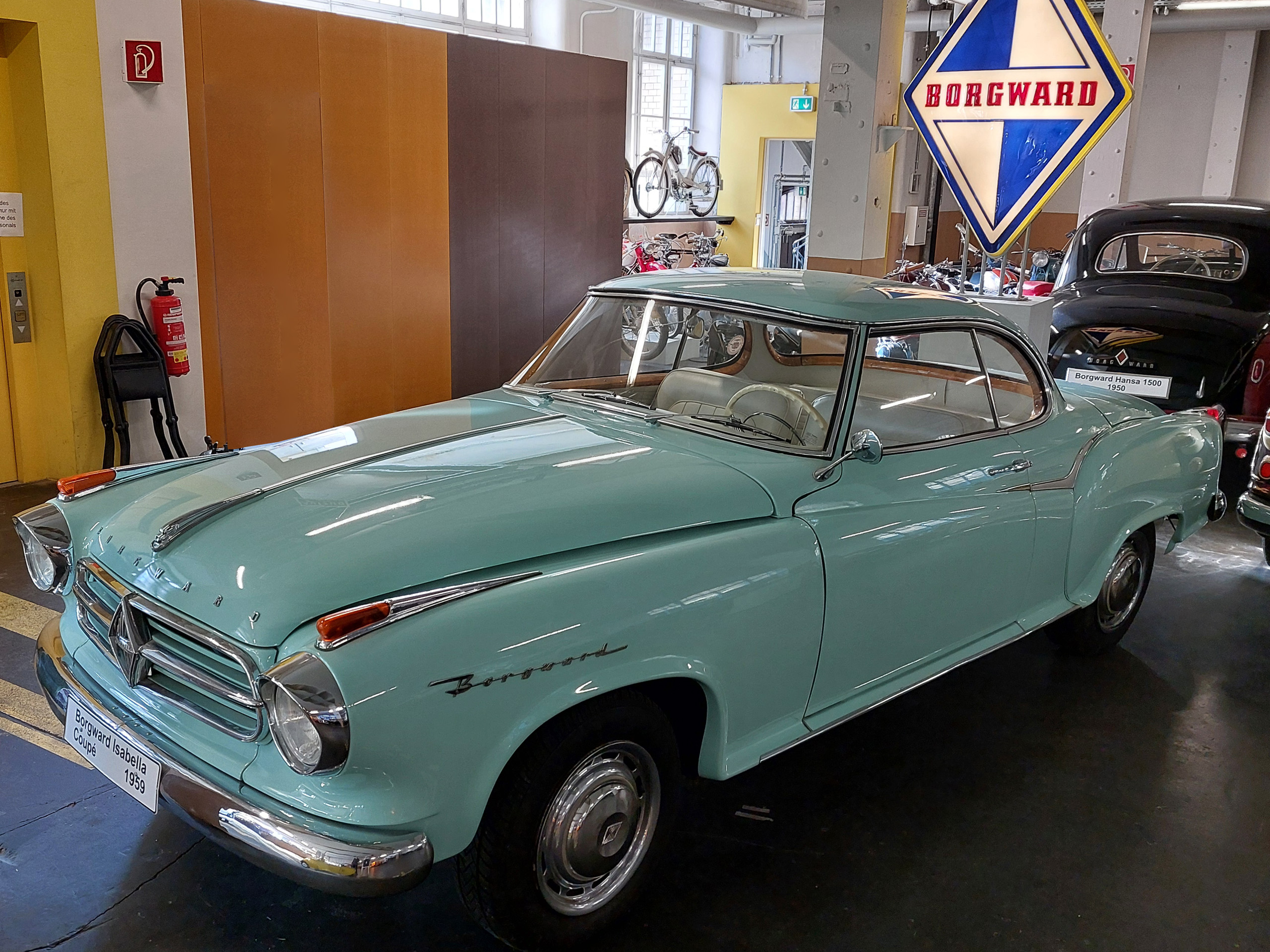Ein mintgrüner Oldtimer der Marke Borgward, Modell Isabella, aus dem Jahr 1958, steht in einem Ausstellungsraum. Das Auto hat geschwungene Linien und Chrom-Applikationen. Im Hintergrund sind weitere historische Fahrzeuge und ein großes Borgward-Schild sichtbar.