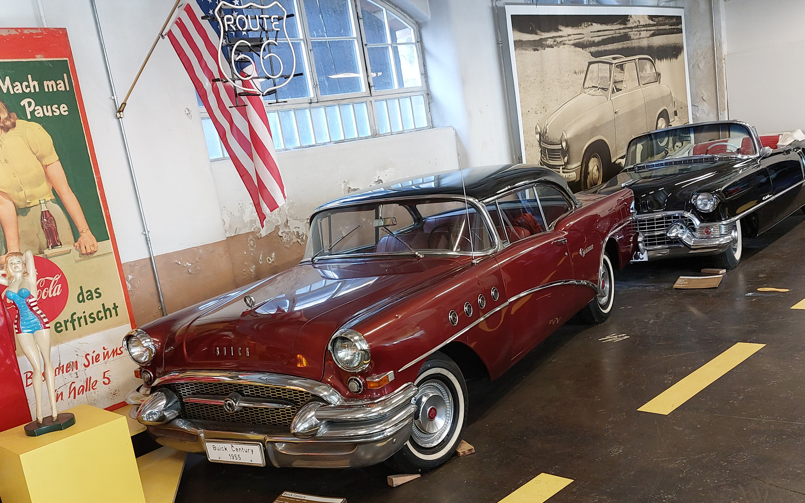 Im Bild sind zwei klassische Autos in einem Museum zu sehen. Das vordere Auto ist ein roter Buick Century von 1955, während das hintere ein schwarzes Cabriolet ist. Im Hintergrund hängen eine amerikanische Flagge und ein Schild mit der Aufschrift „Route 66“.