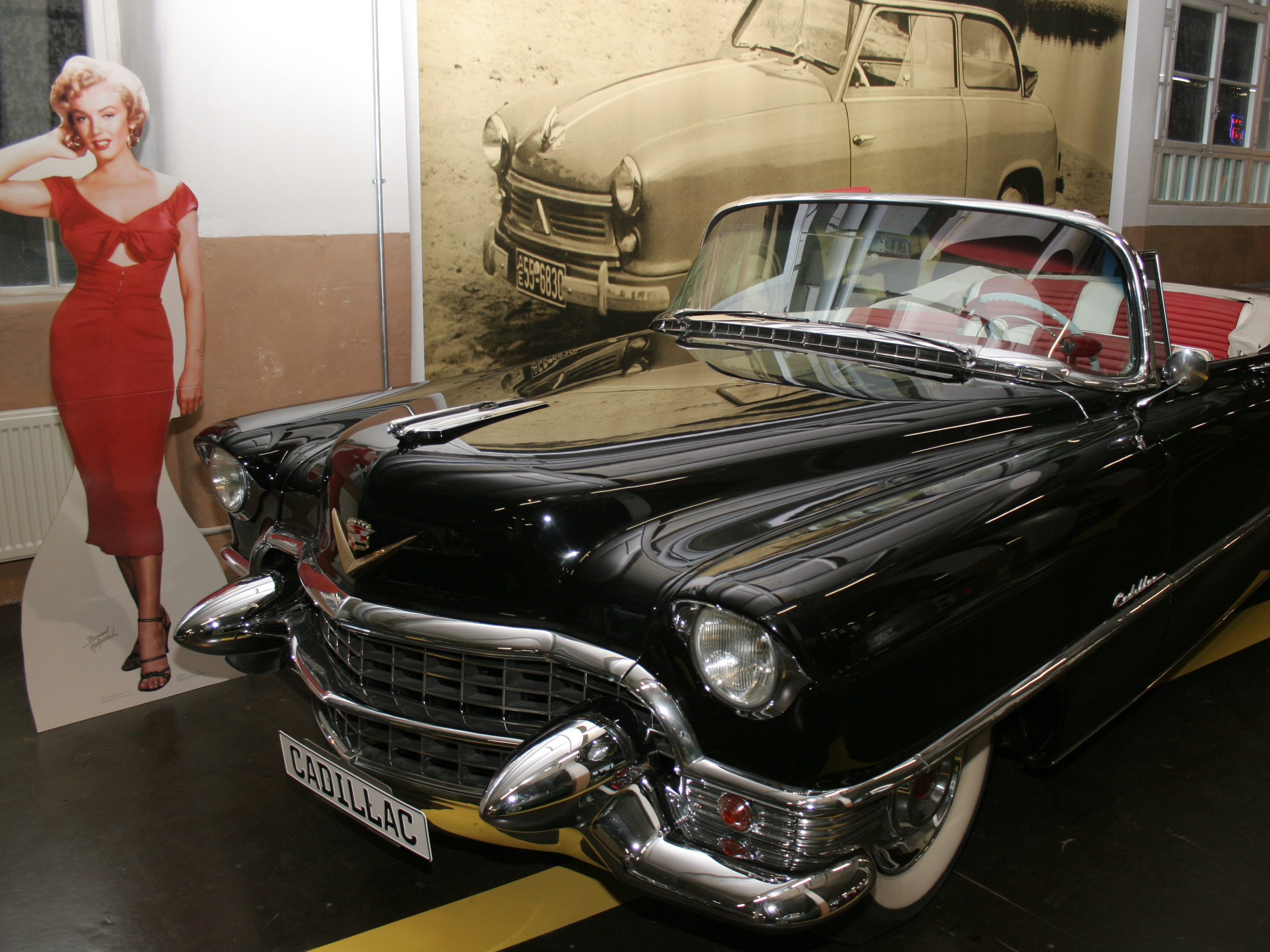 Ein schwarzer Cadillac Cabrio aus den 1950er Jahren steht im Vordergrund. Daneben befindet sich eine lebensgroße Standfigur einer Frau in einem roten Kleid, die eine Hand an ihr Gesicht legt. Im Hintergrund ist ein älteres Auto auf einem Foto zu sehen.