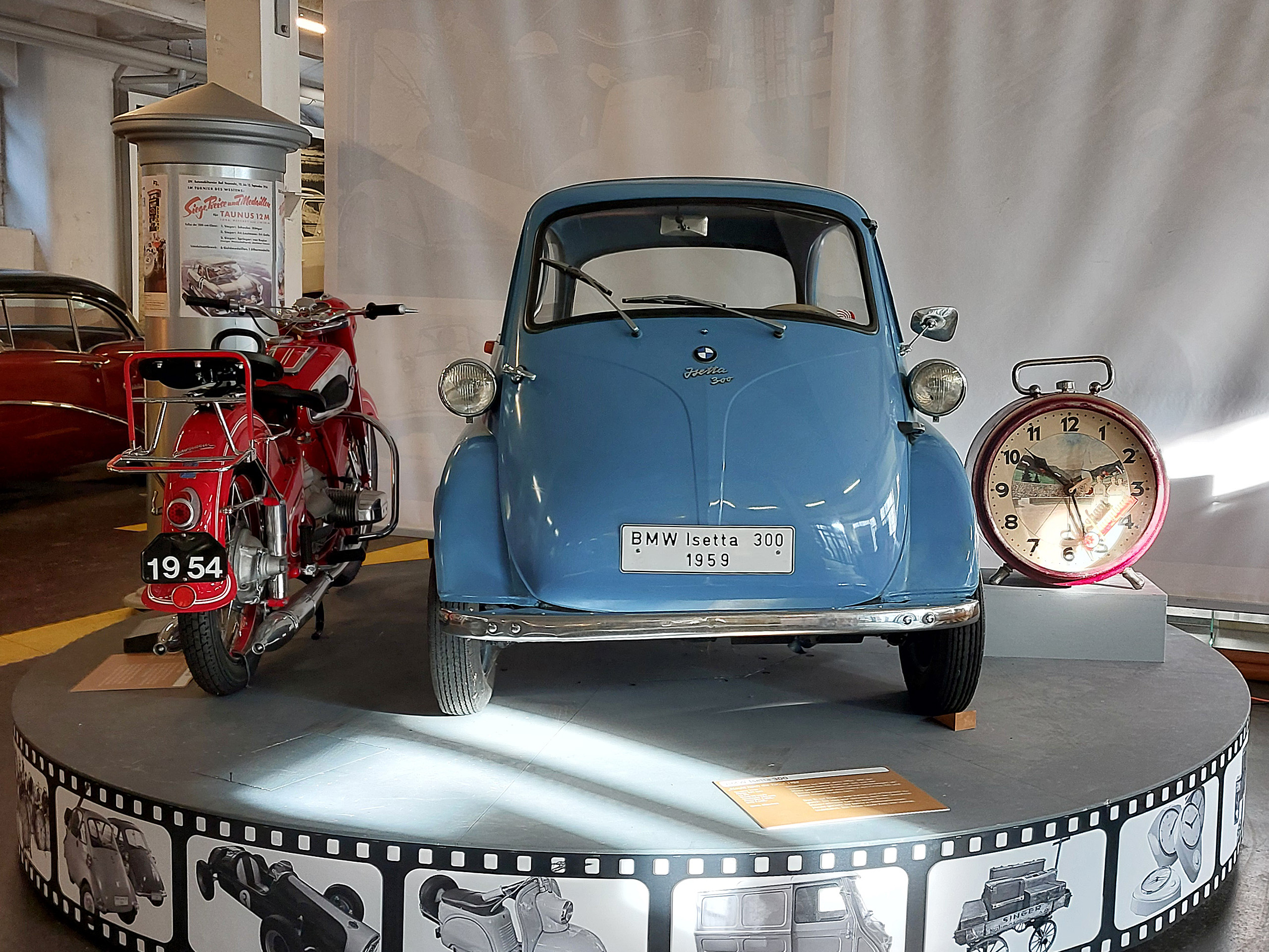 Auf der Ausstellung sind ein rotes Motorrad aus 1954 und ein blauer BMW Isetta 300 aus 1959 auf einem runden Podest zu sehen, begleitet von einer alten, runden Tischuhr. Der Hintergrund ist hell und neutral, um die Fahrzeuge hervorzuheben.