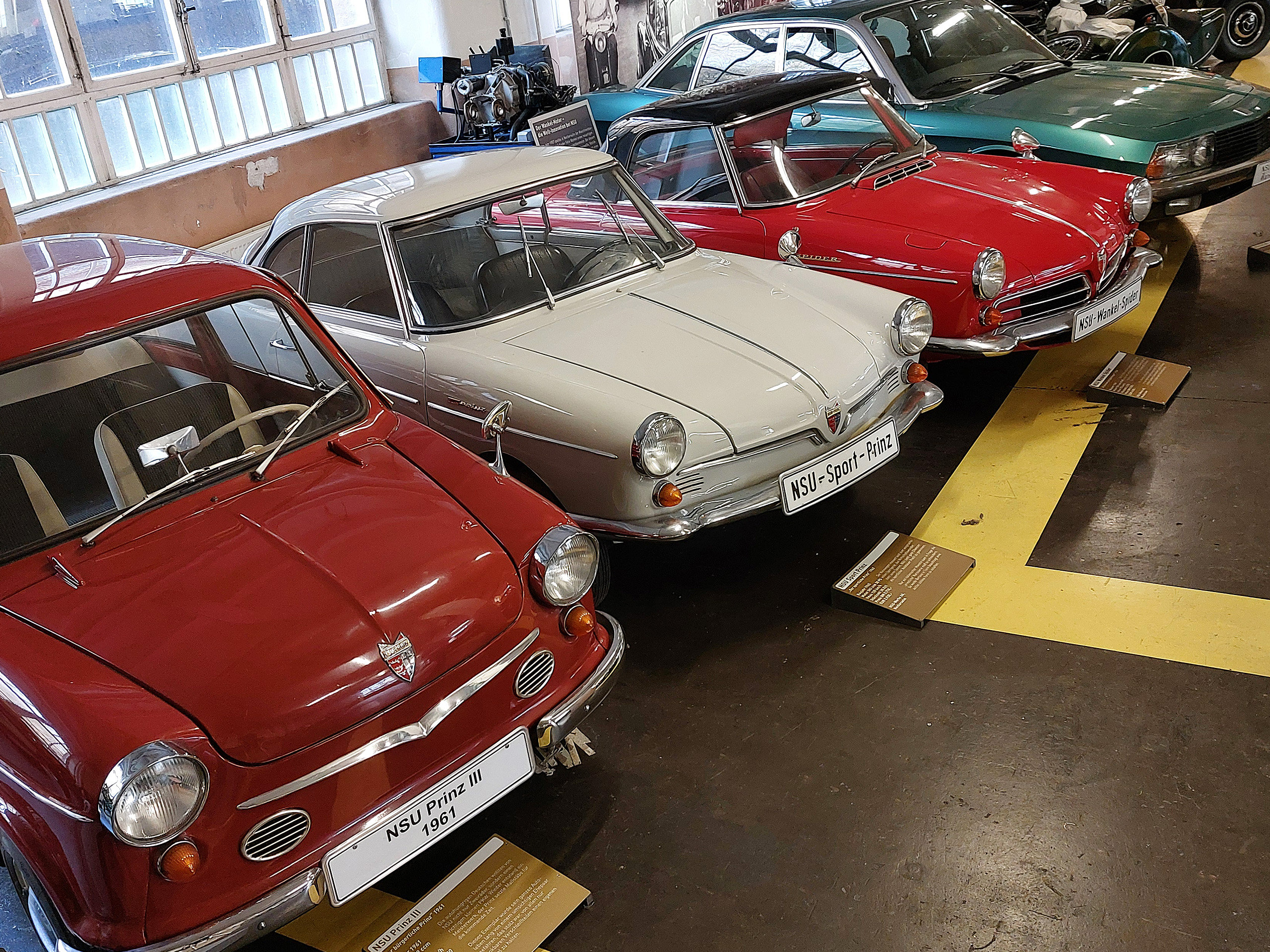Im Bild sind mehrere klassische Automobile in einem Museum zu sehen. Im Vordergrund steht ein roter NSU Prinz III von 1961, daneben ein weißer NSU Sport-Prinz. Im Hintergrund sind weitere alte Autos zu erkennen. Die Fahrzeuge sind in einem geschlossenen Raum ausgestellt.