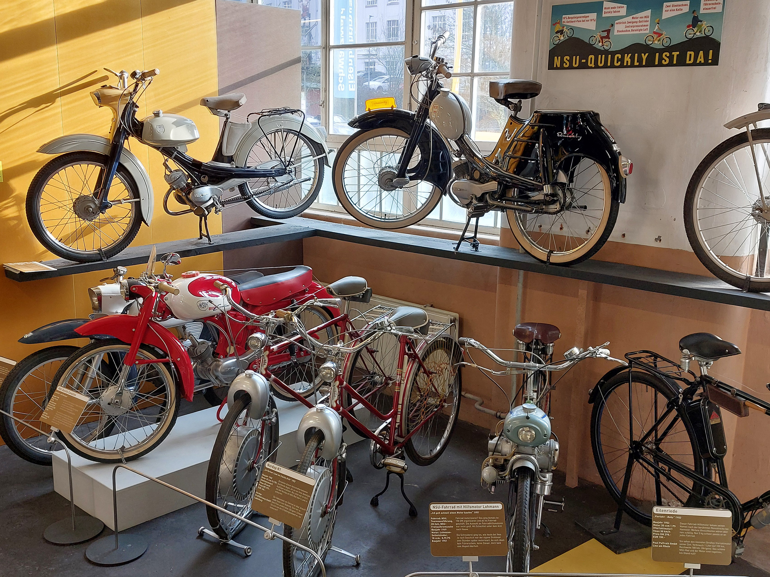 Eine Ausstellung von alten Fahrrädern und Mopeds. Im Vordergrund stehen rote und silberne Modelle, im Hintergrund sind weitere Fahrräder auf Regalen angeordnet. Ein gelbes Licht fällt durch ein Fenster und beleuchtet die Fahrzeuge, während ein Schild mit der Aufschrift "NSU-Quickly ist da!" im Hintergrund sichtbar ist.