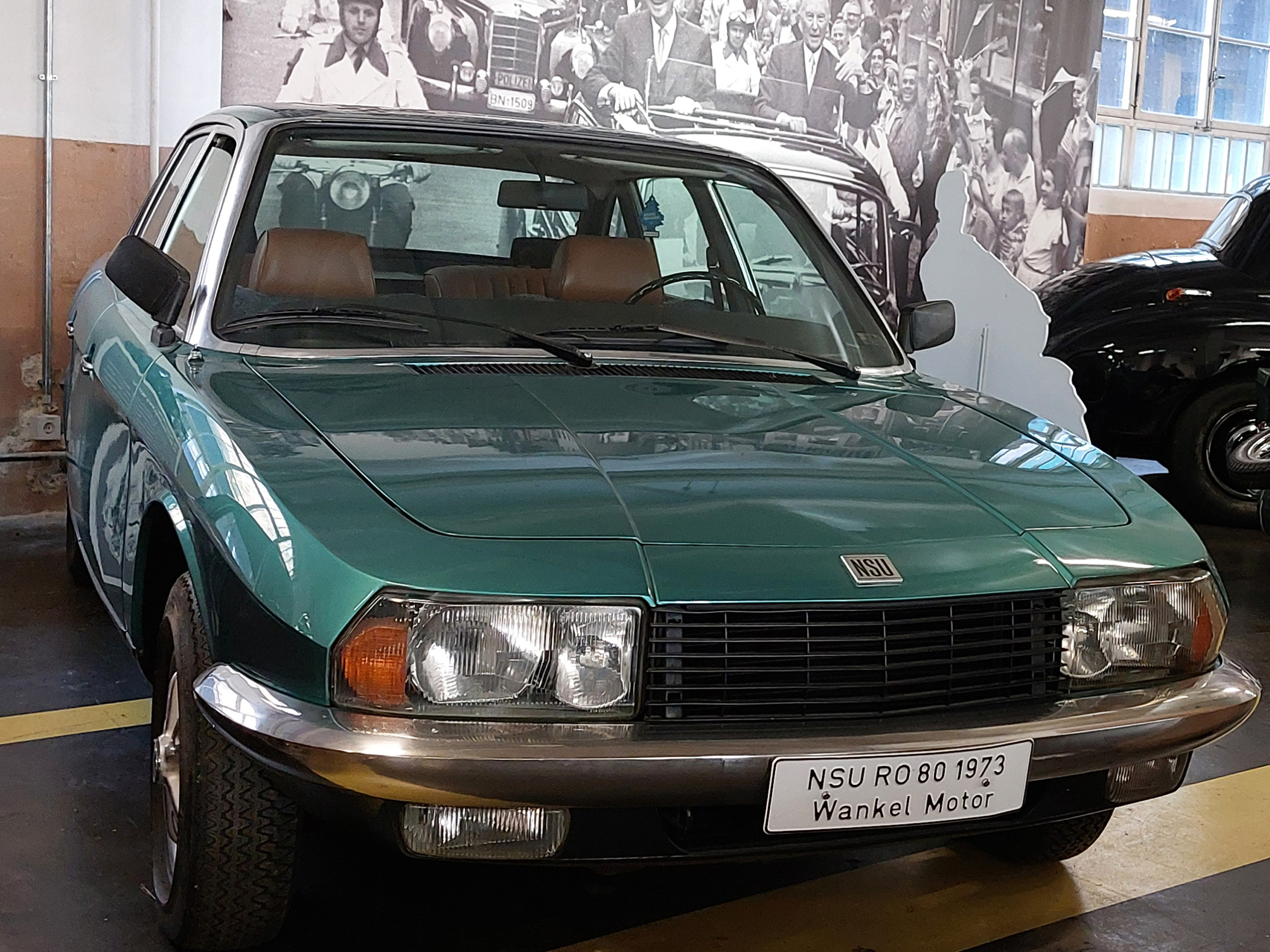 Ein grüner NSU RO 80 aus dem Jahr 1973 steht auf einem Ausstellungsboden. Das Auto hat eine elegante Karosserie mit einem markanten Kühlergrill und großen Scheinwerfern. Ein Schild zeigt den Modellnamen und den Hinweis auf den Wankelmotor.