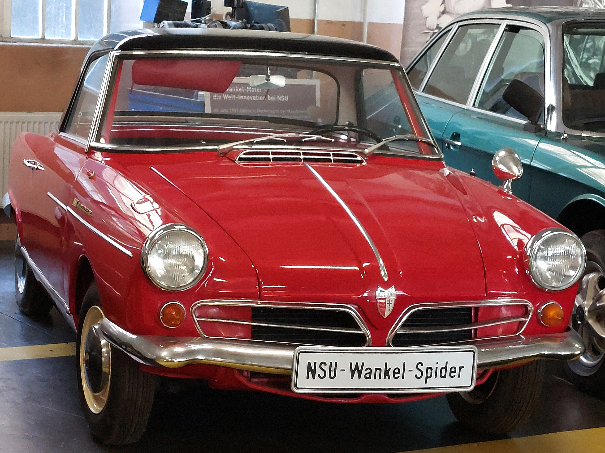 Ein roter NSU Wankel-Spider steht in einer Ausstellung. Das Auto hat einen runden Frontscheinwerfer, einen chromierten Kühlergrill und eine sportliche Form. Es ist ein klassisches Modell mit einem zweifarbigen Design, das seinen Retro-Charme ausstrahlt.