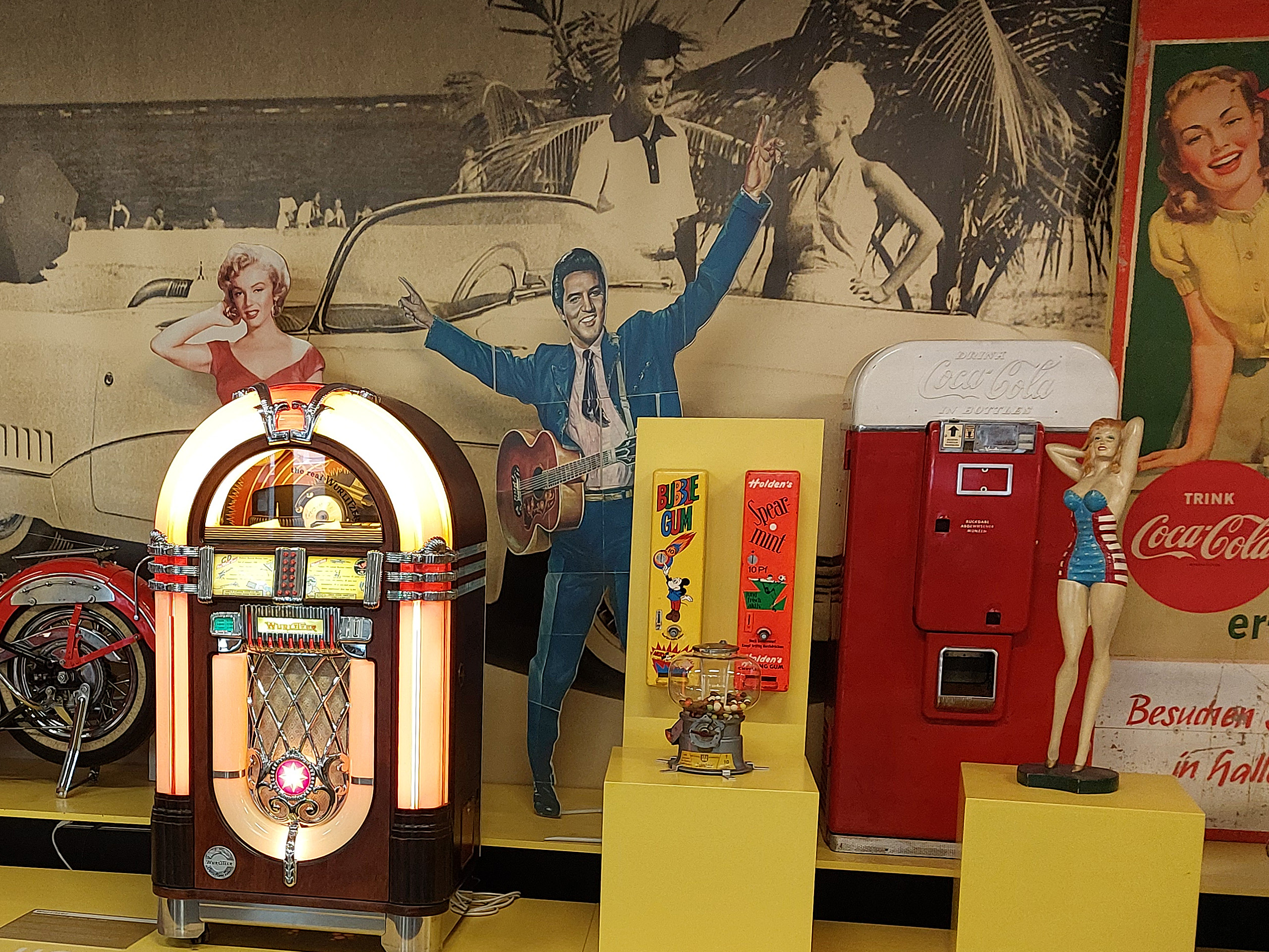 Eine vintage Musikbox steht im Vordergrund, umgeben von nostalgischen Objekten wie einem Cola-Automaten und bunten Figuren. Im Hintergrund sieht man stilisierte Darstellungen berühmter Personen aus der Zeit, die eine sommerliche Strandatmosphäre vermitteln.