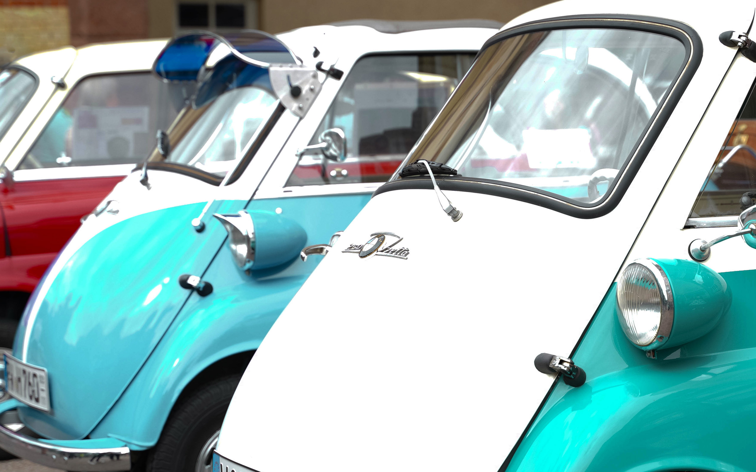 Drei farbenfrohe Fahrzeuge stehen nebeneinander. Die vorderen Fahrzeuge sind in Türkis mit weißem Dach und glänzenden Chromdetails. Im Hintergrund sind weitere Fahrzeuge in verschiedenen Farben zu erkennen. Die Vorstellung vermittelt einen nostalgischen Charme und eine lebendige, freundliche Atmosphäre.