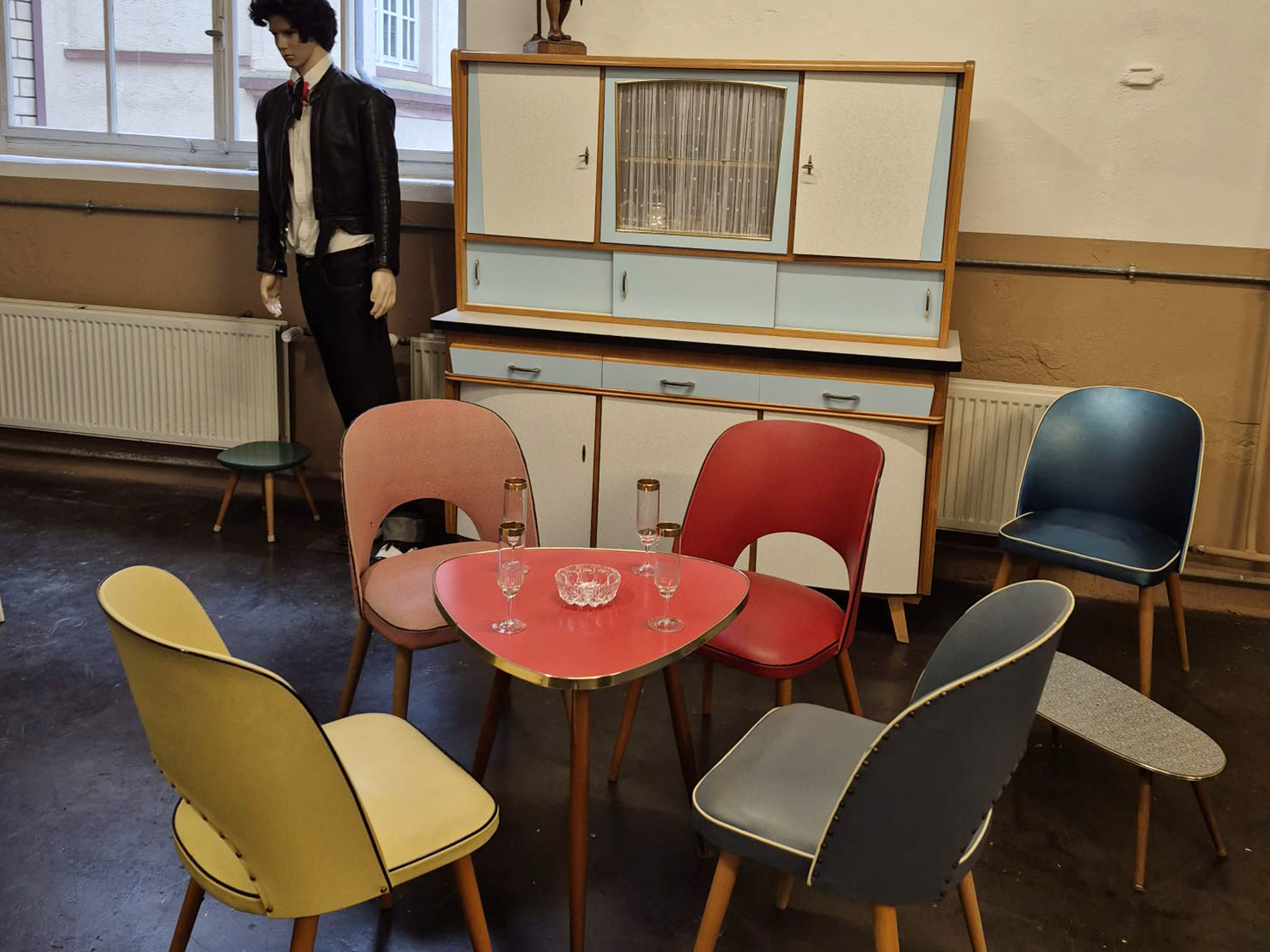 Ein retro-moderner Essbereich mit einem runden Tisch in Rot und sechs bunten Stühlen in unterschiedlichen Farben (gelb, pink, rot, blau). Auf dem Tisch stehen zwei Sektgläser und eine kleine Glasvase. Im Hintergrund steht ein Möbelstück mit Glasfronten und Schubladen.