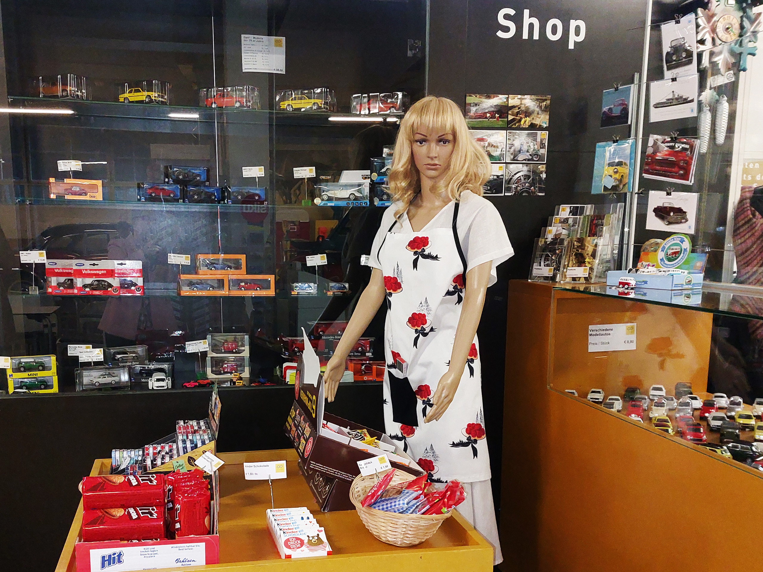 Ein Schaufenster mit einer Schaufensterpuppe in einem weißen Kleid mit roten Blumen. Hinter der Puppe steht ein Regal mit verschiedenen Modellautos und Postkarten. Auf einem Tisch im Vordergrund liegen Süßigkeiten und kleine Artikel zum Verkauf, während ein Schild mit dem Wort "Shop" über der Szene hängt.
