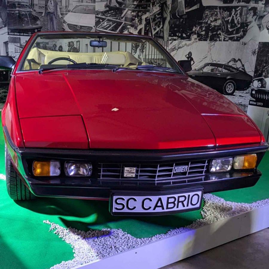 Ein rotes Cabriolet steht frontal im Bild. Es hat eine elegante Form mit klaren Linien und einem schwarzen Kühlergrill. Das Auto steht auf grünem Teppich, der mit kleinen Steinen dekoriert ist. Im Hintergrund sind verschwommene Bilder von weiteren Autos zu sehen.