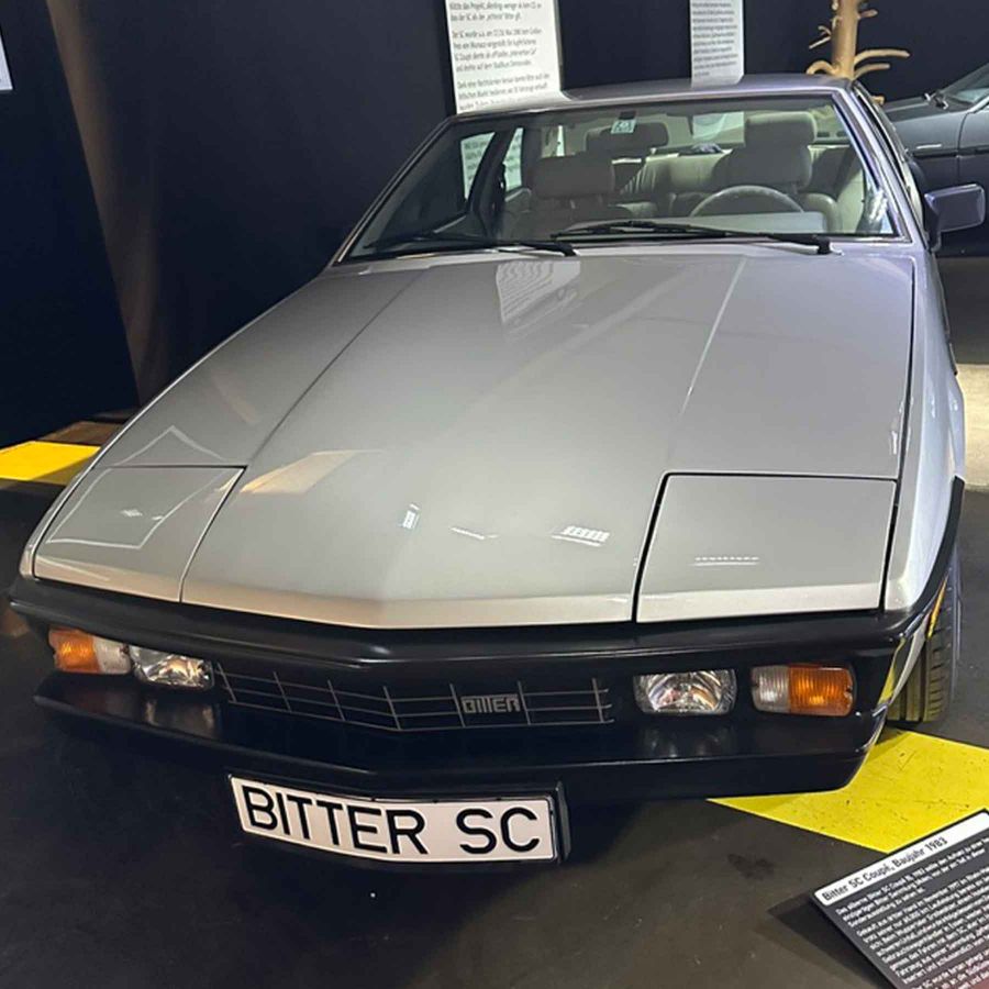 Ein silberner Bitter SC Sportwagen steht frontal im Bild. Der Wagen hat ein markantes, flaches Design mit einer breiten Motorhaube und einem schwarzen Kühlergrill. Die Scheinwerfer sind gut sichtbar, und ein Nummernschild mit der Aufschrift "BITTER SC" ist angebracht.