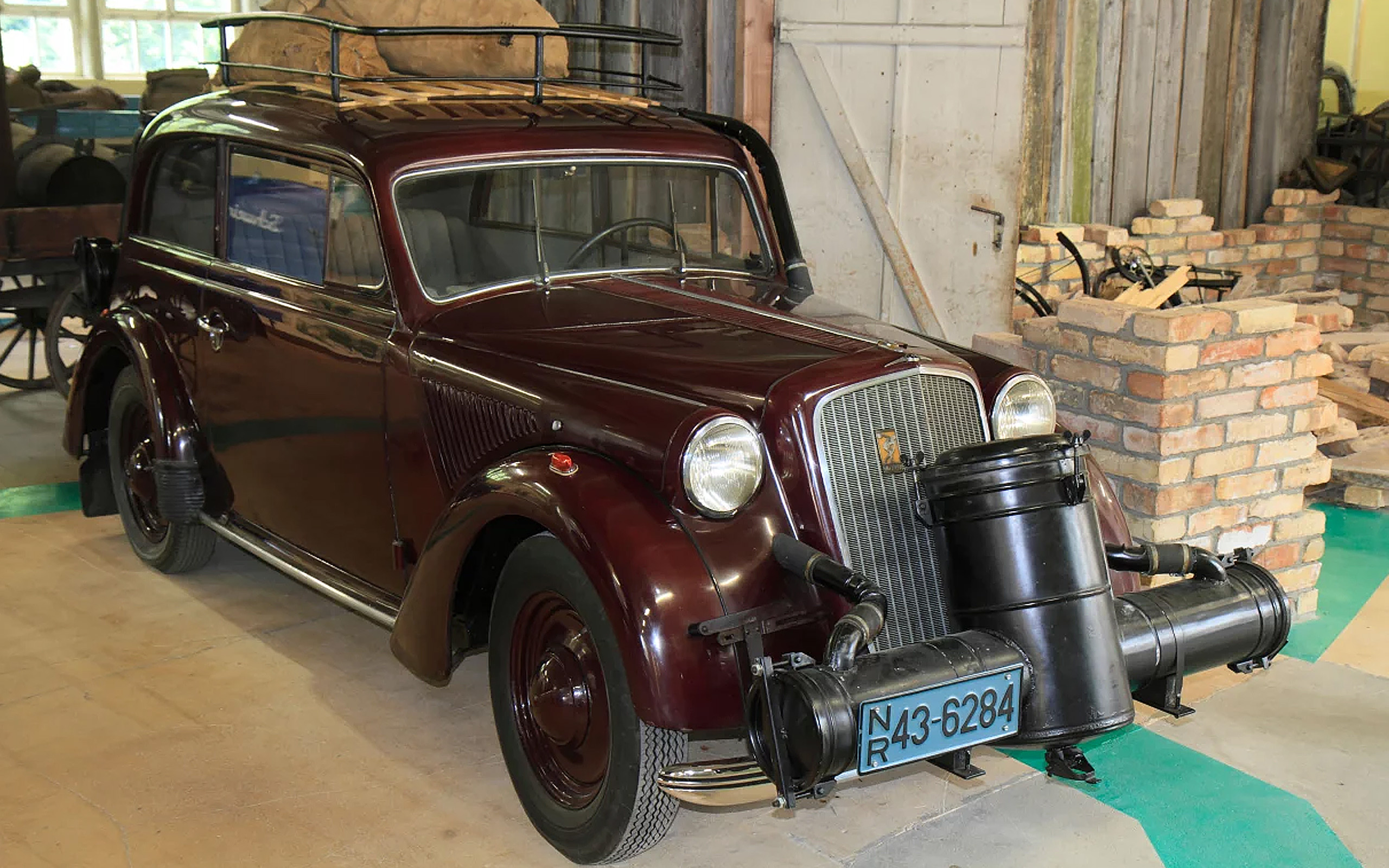 Ein roter Oldtimer mit zwei runden Scheinwerfern und einem markanten Kühlergrill steht in einer Werkstatt. Auf dem Dach befindet sich ein Gepäckträger mit einem großen Holzsack. Der Wagen ist bis in die kleinsten Details gut erhalten und vermittelt ein nostalgisches Gefühl.