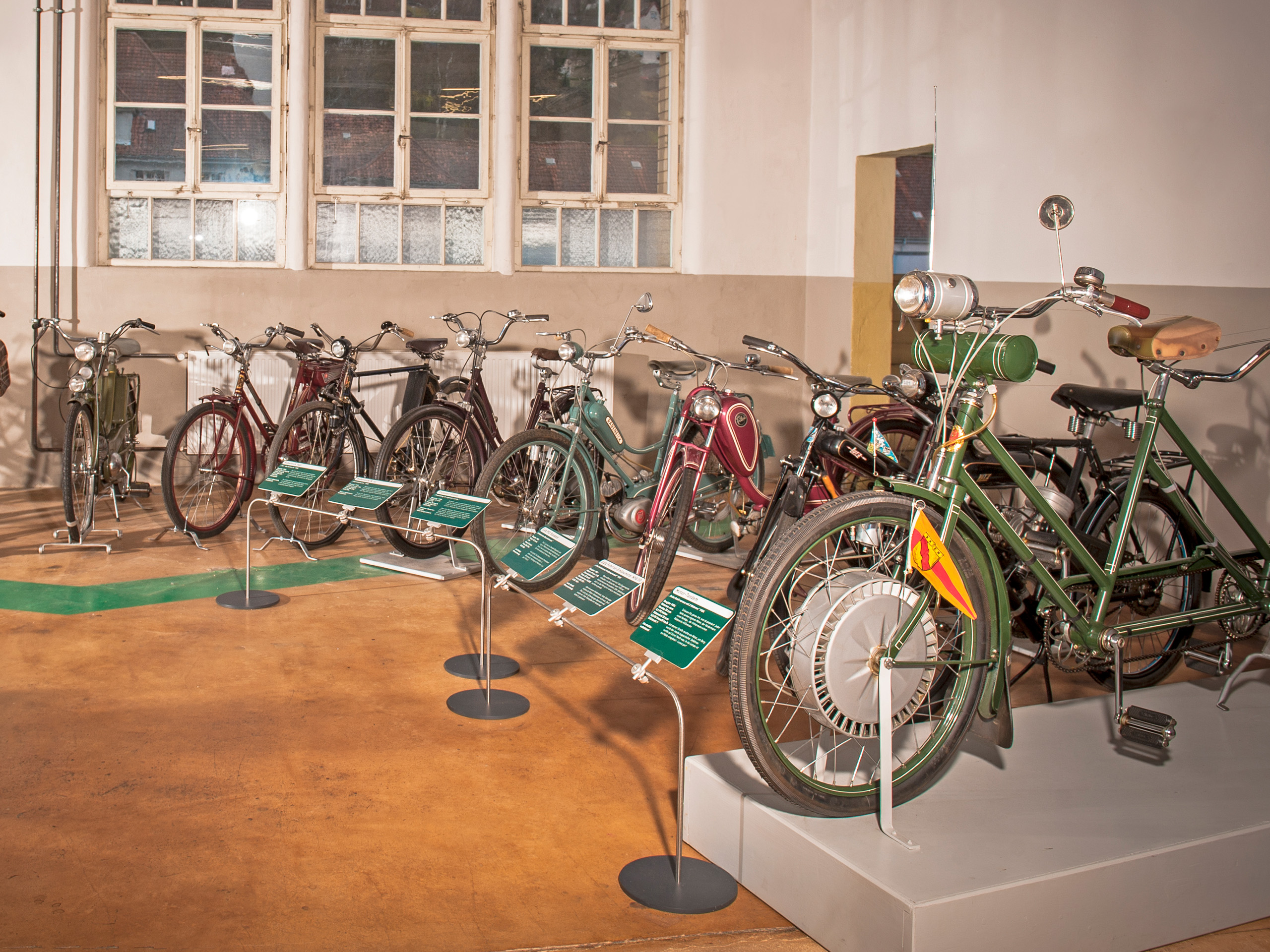 Das Bild zeigt eine Ausstellung mit mehreren Fahrrädern unterschiedlichen Designs in einem Raum. Jedes Fahrrad steht auf einem Ständer, und einige haben Informationstafeln daneben. Die Fahrräder sind in verschiedenen Farben, wie Grün, Rot und Blau, gehalten.