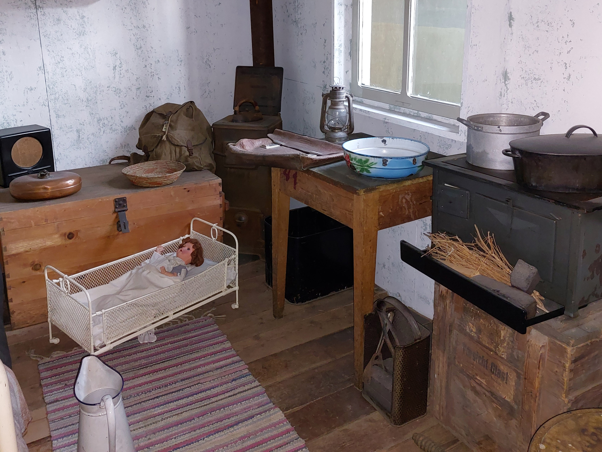 Das Bild zeigt eine nostalgische Küche mit einem rustikalen Holztisch, einem alten Herd und einem kleinen Korb mit einer liegenden Puppe in einem Bettchen. Daneben sind verschiedene Küchenutensilien und ein Fenster, das natürliches Licht hereinlässt. Der Boden ist mit einem gestreiften Teppich bedeckt.