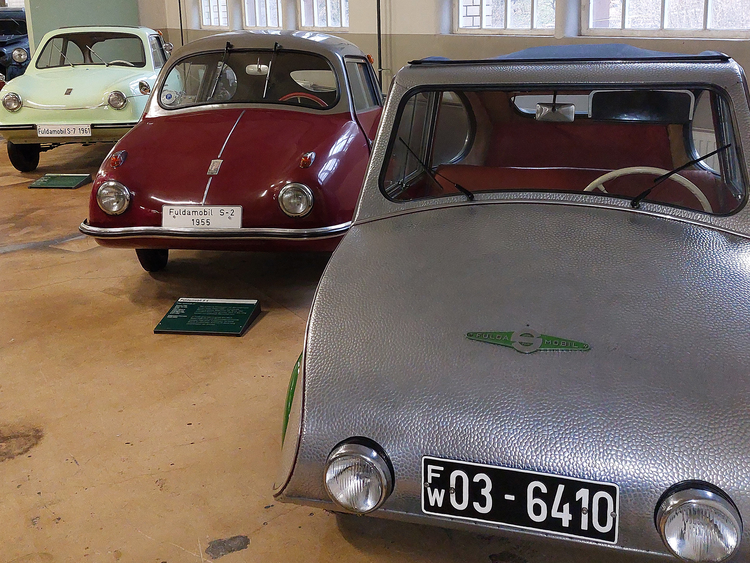 Drei historische Kleinstwagen sind in einem Ausstellungsraum zu sehen. Der vorderste Wagen ist silbern mit einer ovalen Karosserie und zwei runden Scheinwerfern. Im Hintergrund stehen ein roter und ein mintfarbener Wagen, beide ebenfalls kompakt und nostalgisch gestaltet.