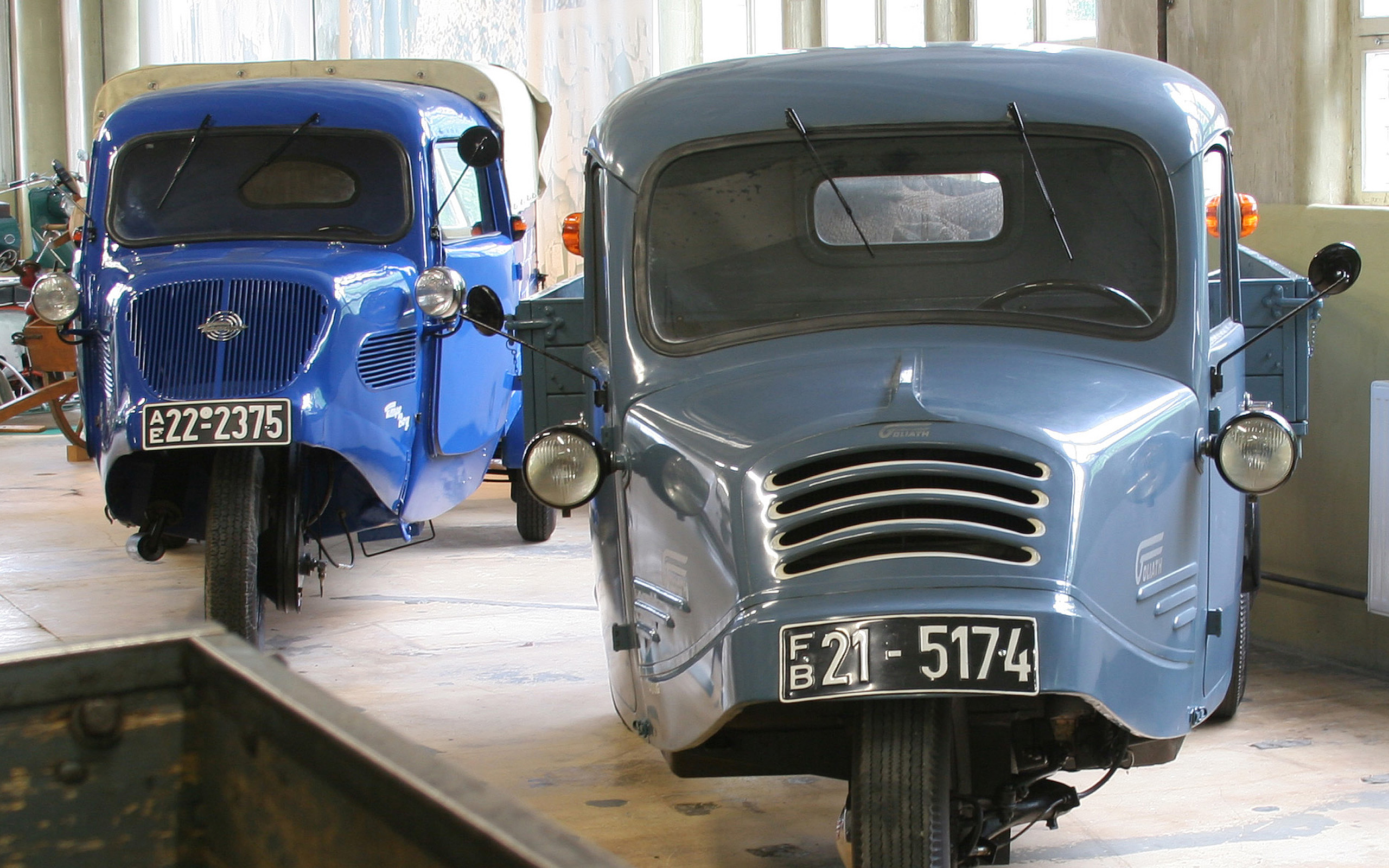 Das Bild zeigt zwei historische Dreiradfahrzeuge in einem Museum. Das linke Fahrzeug ist blau und hat einen runden Kühlergrill. Das rechte Fahrzeug ist grau und hat einen charakteristischen Frontgrill mit horizontalen Lücken. Beide Fahrzeuge stehen nebeneinander in einem beleuchteten Raum.