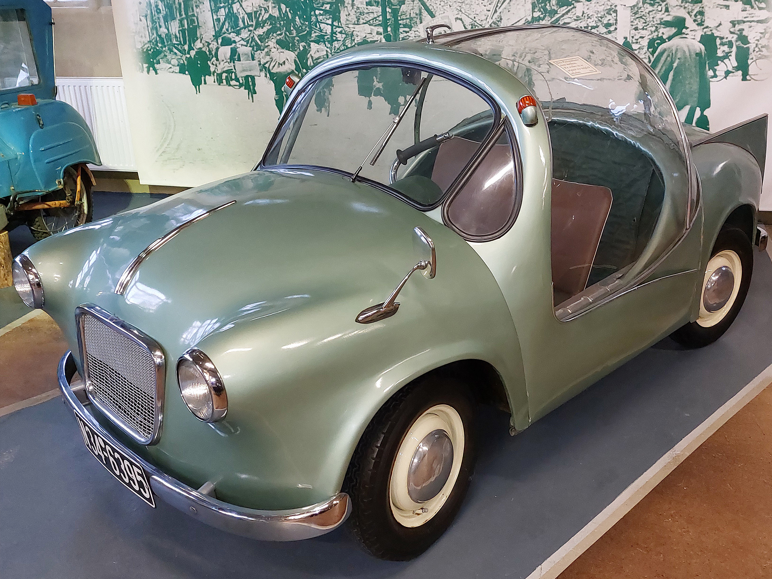 Ein kleiner, rundlicher, mintgrüner Oldtimer mit transparentem Dach und offenen Seiten. Die Front hat einen rechteckigen Kühlergrill und runde Scheinwerfer. Das Auto steht auf einem hellen Boden, umgeben von einer beschränkten, historischen Ausstellung.