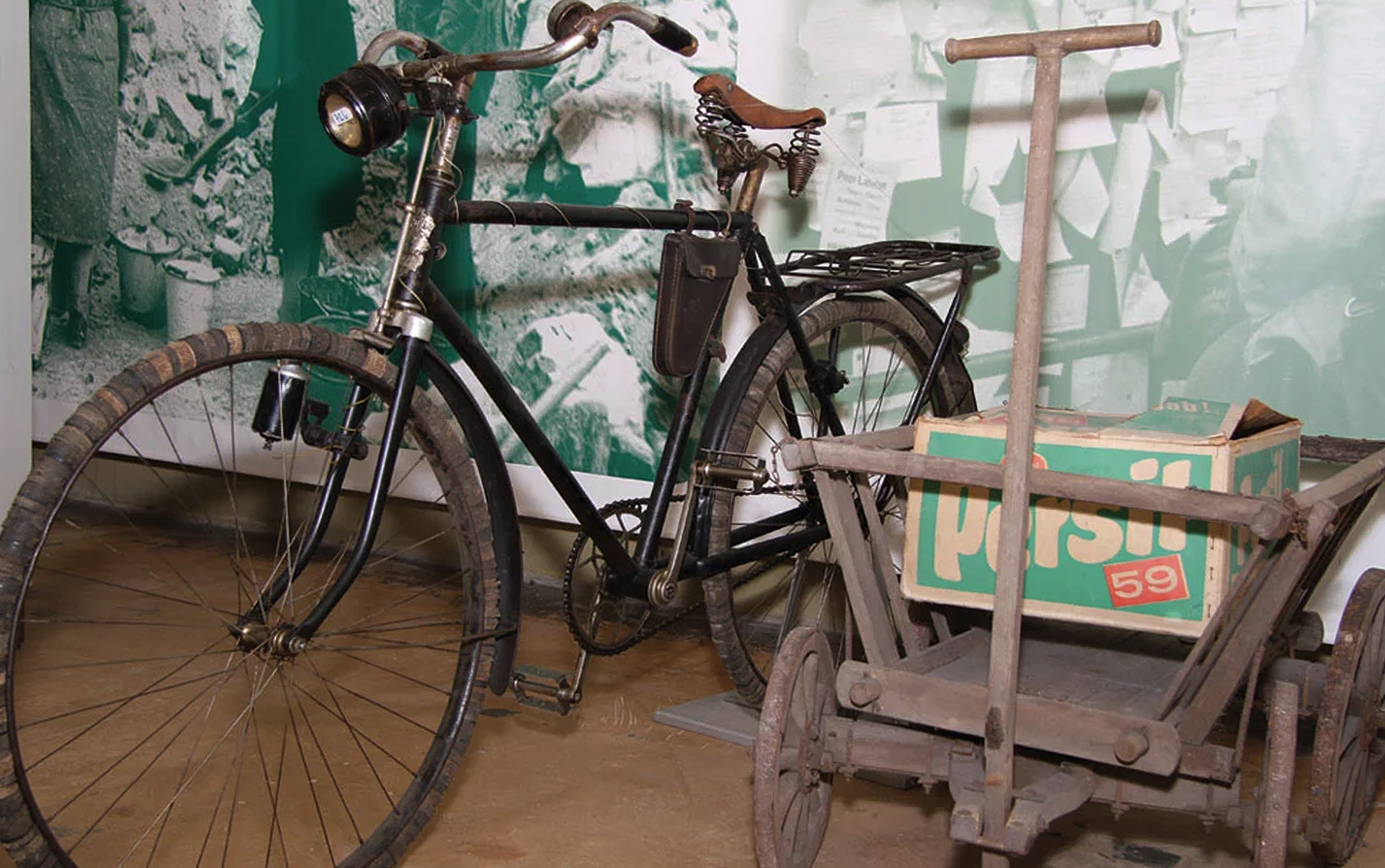 Ein altes schwarz lackiertes Fahrrad mit einem Korb auf dem Gepäckträger steht neben einem Holzwagen mit einer Kiste. Der Wagen hat zwei Räder und einen langen Griff. Im Hintergrund sind unscharfe grüne Wandbilder sichtbar.