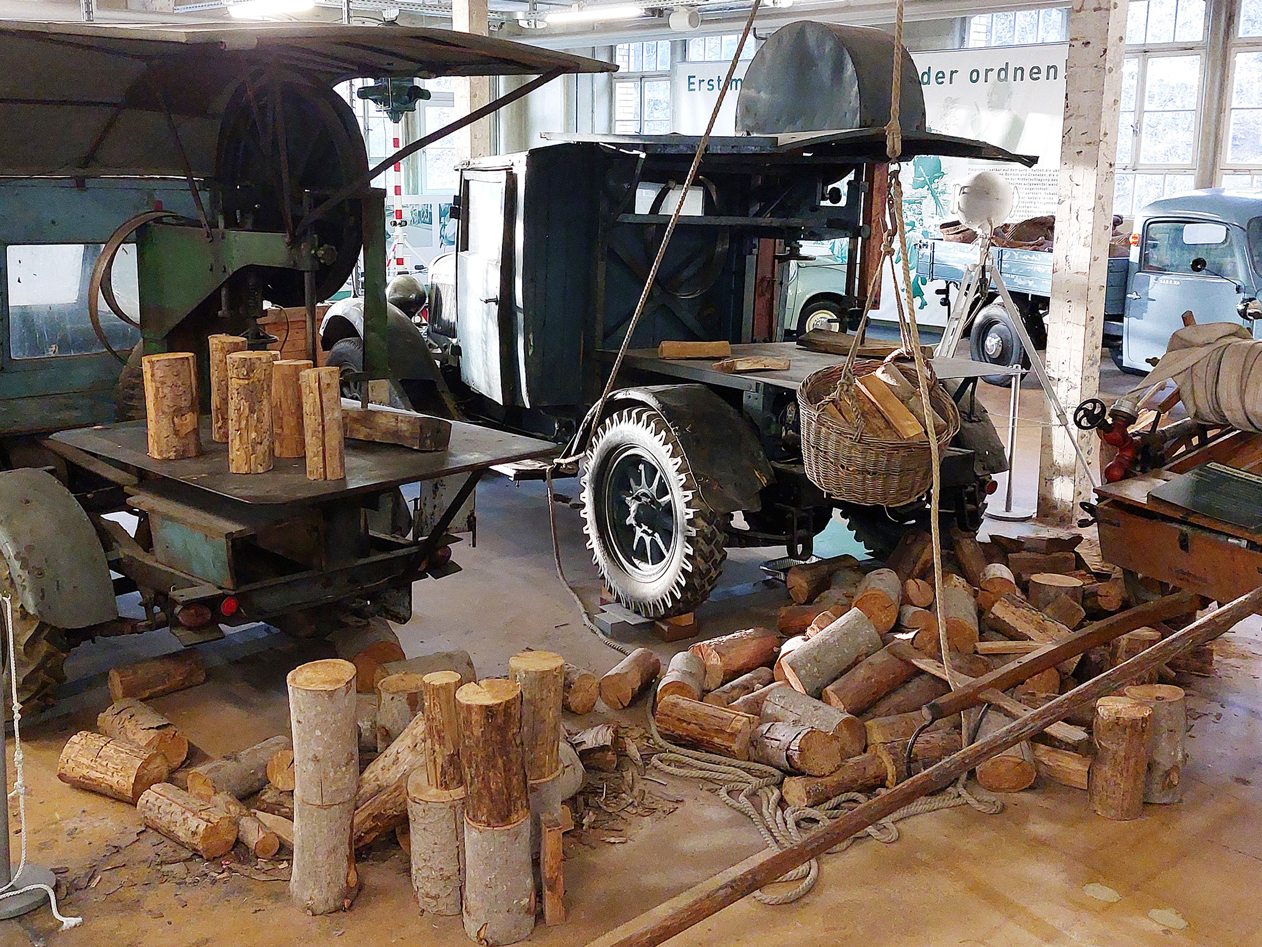 Eine alte Holzverarbeitungsmaschine steht in einem Raum, umgeben von mehreren Holzstämmen und -stücken. Ein gelbes Fahrzeug mit einem besonderen Design ist im Hintergrund sichtbar. Der Boden ist mit zerlegtem Holz und Seilen bedeckt.