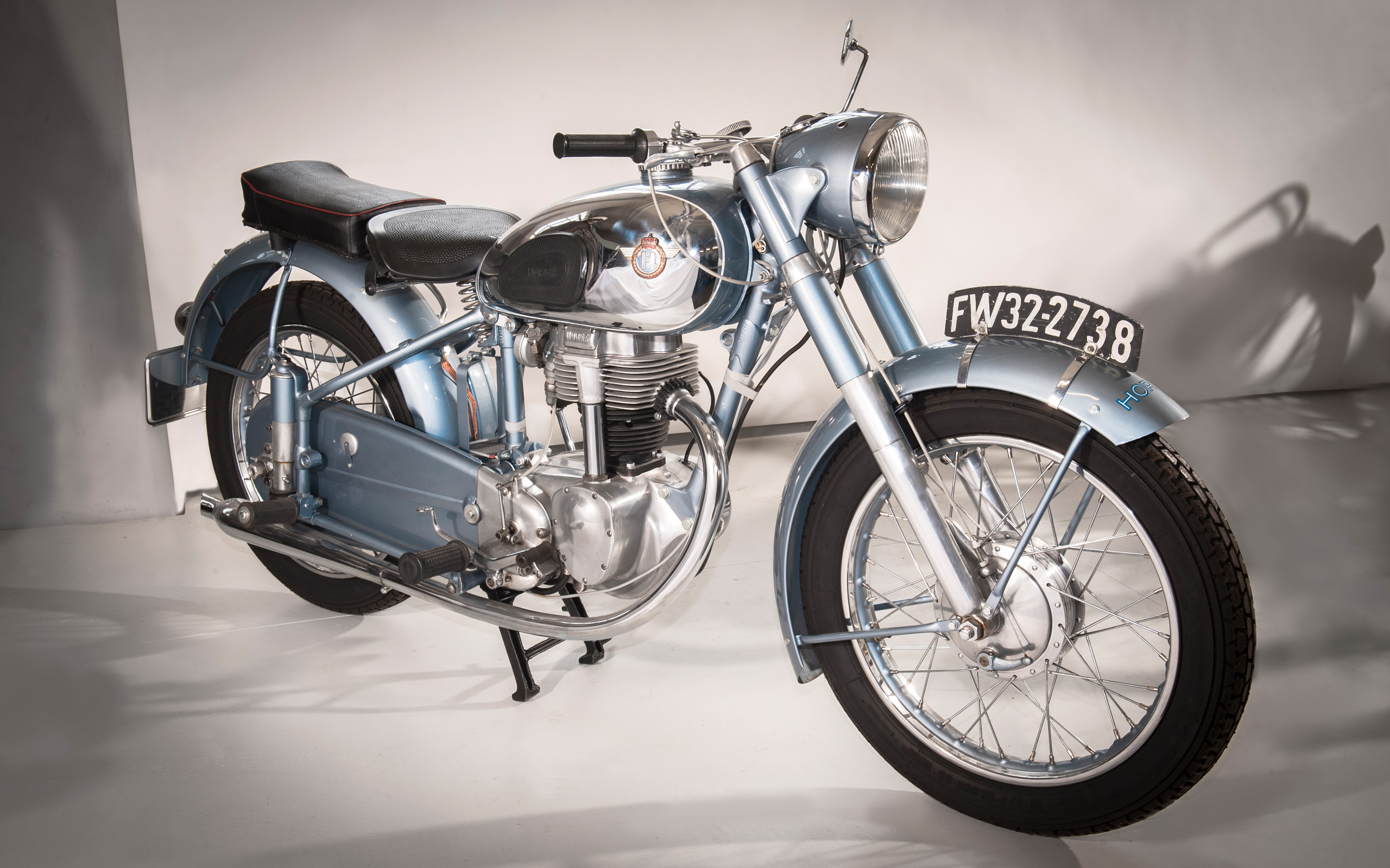 Ein klassisches Motorrad in hellem Blau mit chromfarbenen Details. Der Tank hat ein Logo, und das Motorrad steht auf einer weißen Fläche. Es hat einen schwarzen Sitz und eine glänzende Scheinwerferverkleidung. Im Hintergrund sind neutrale Farben zu sehen.