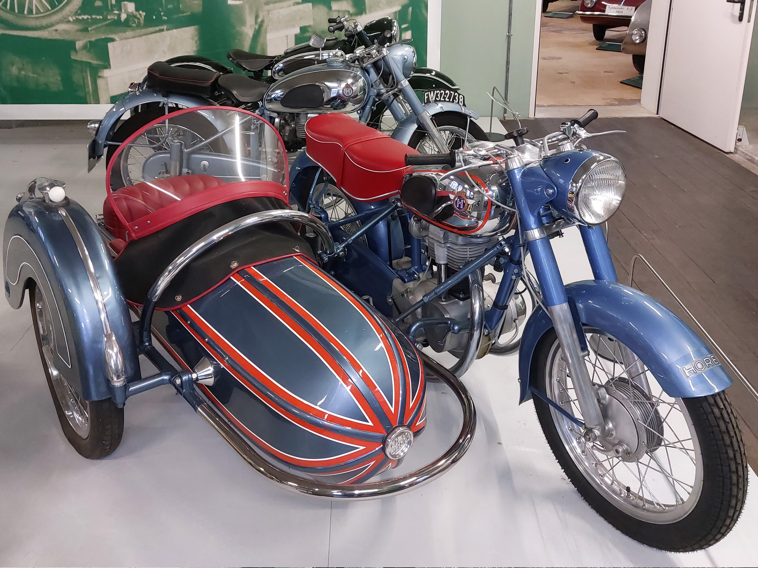 Ein klassisches Motorrad mit einem blauen Seitenwagen, der rot gepolstert ist. Das Motorrad hat einen glänzenden Chromscheinwerfer und zeigt dekorative Streifen auf dem Seitenwagen. Im Hintergrund sind weitere Motorräder zu sehen, die eine nostalgische Sammlung bilden.