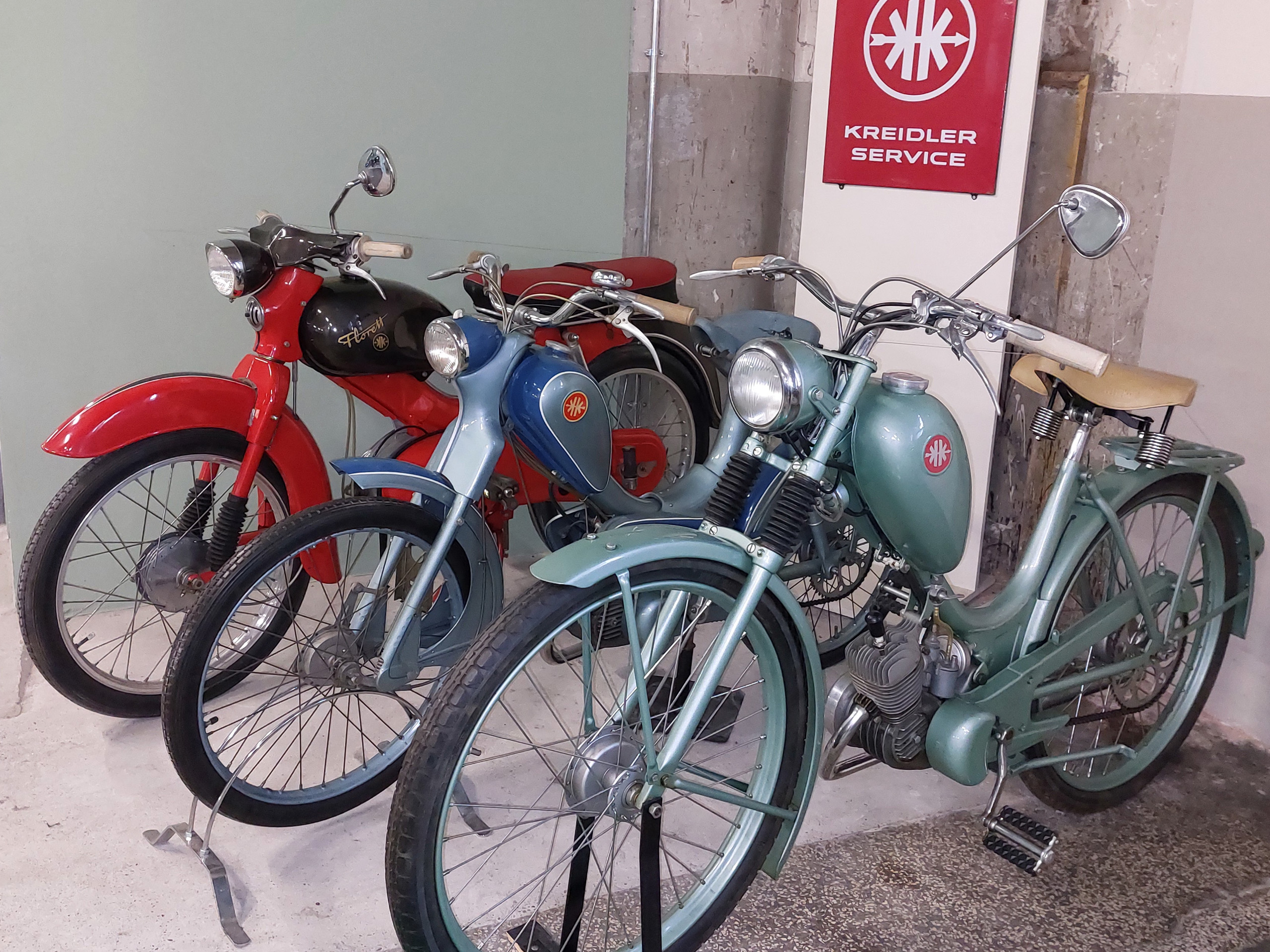 Drei klassische Fahrräder stehen in einer Garage. Ein rotes, ein blaues und ein mintgrünes Fahrrad sind sichtbar. Im Hintergrund hängt ein Schild mit dem Schriftzug „KREIDLER SERVICE“. Die Fahrräder haben retro-Design und sind gut erhalten.